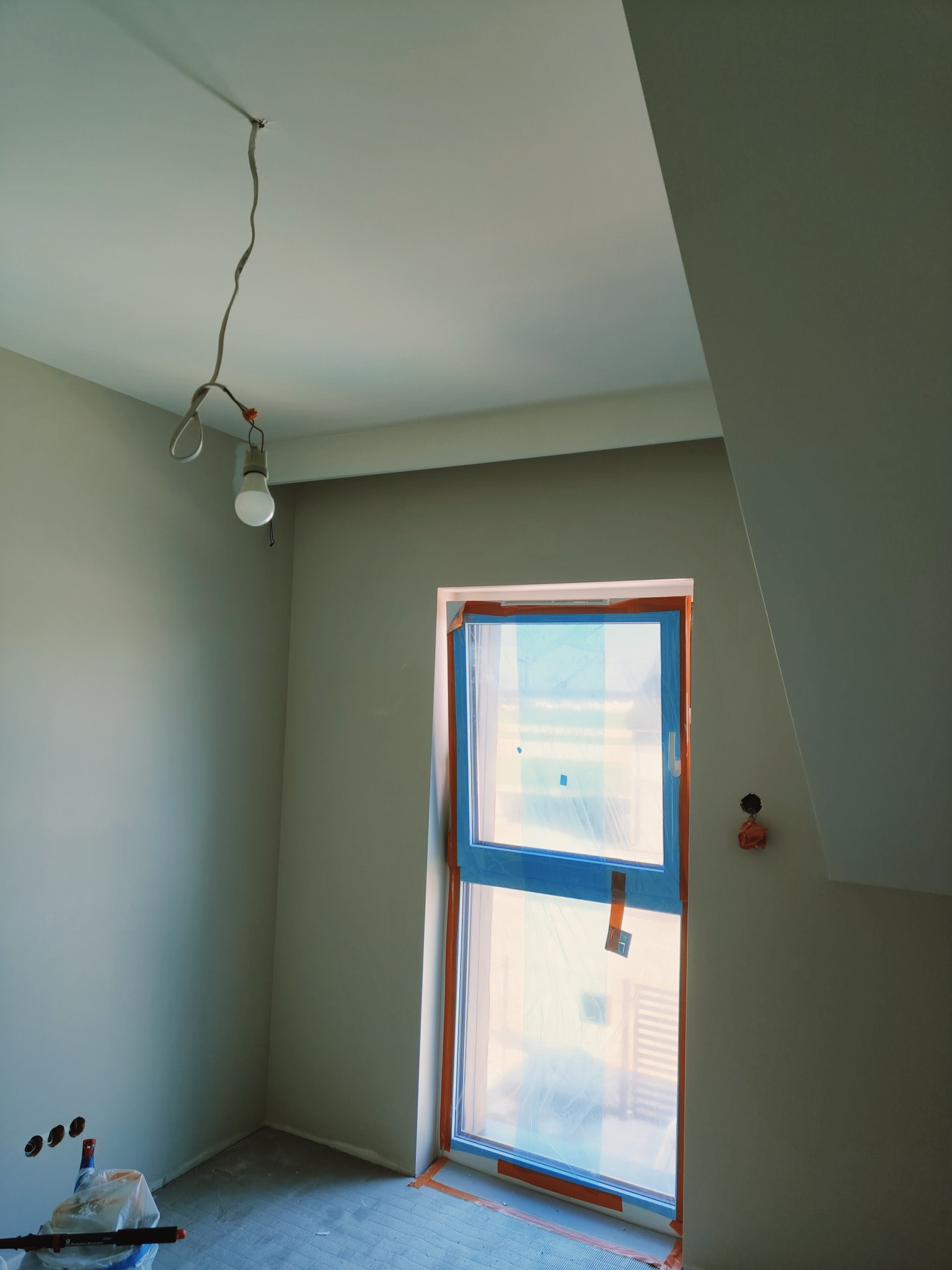 Pokój w trakcie prac wykończeniowych w Obornikach: ściany przygotowane do malowania, okno zabezpieczone folią, widoczne narzędzia malarskie i prowizoryczne oświetlenie.
