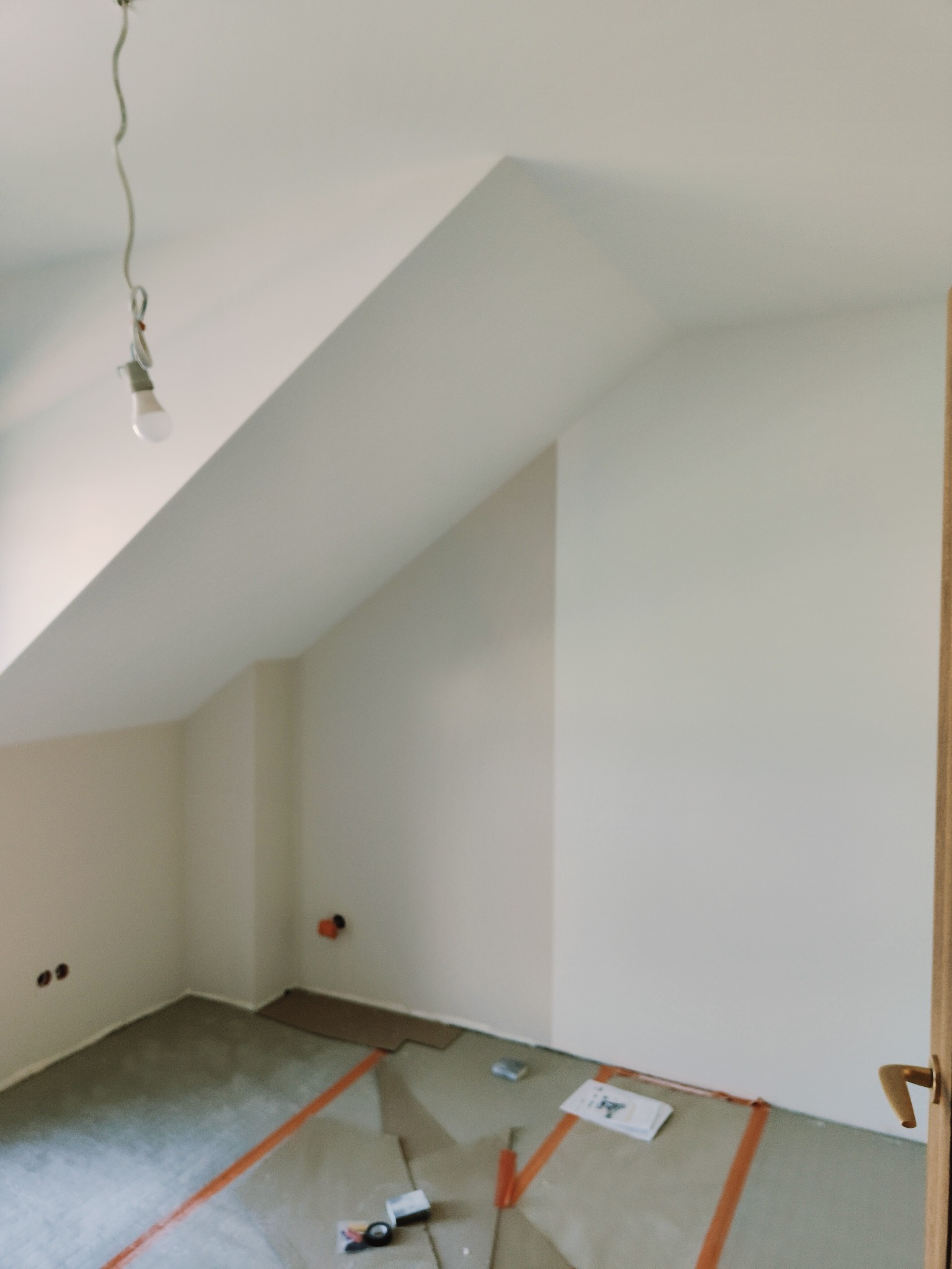 Poddasze w trakcie prac wykończeniowych: ściany pomalowane na biało, widoczne ślady po szpachlowaniu na podłodze zabezpieczonej folią, surowa żarówka zwisa z sufitu.