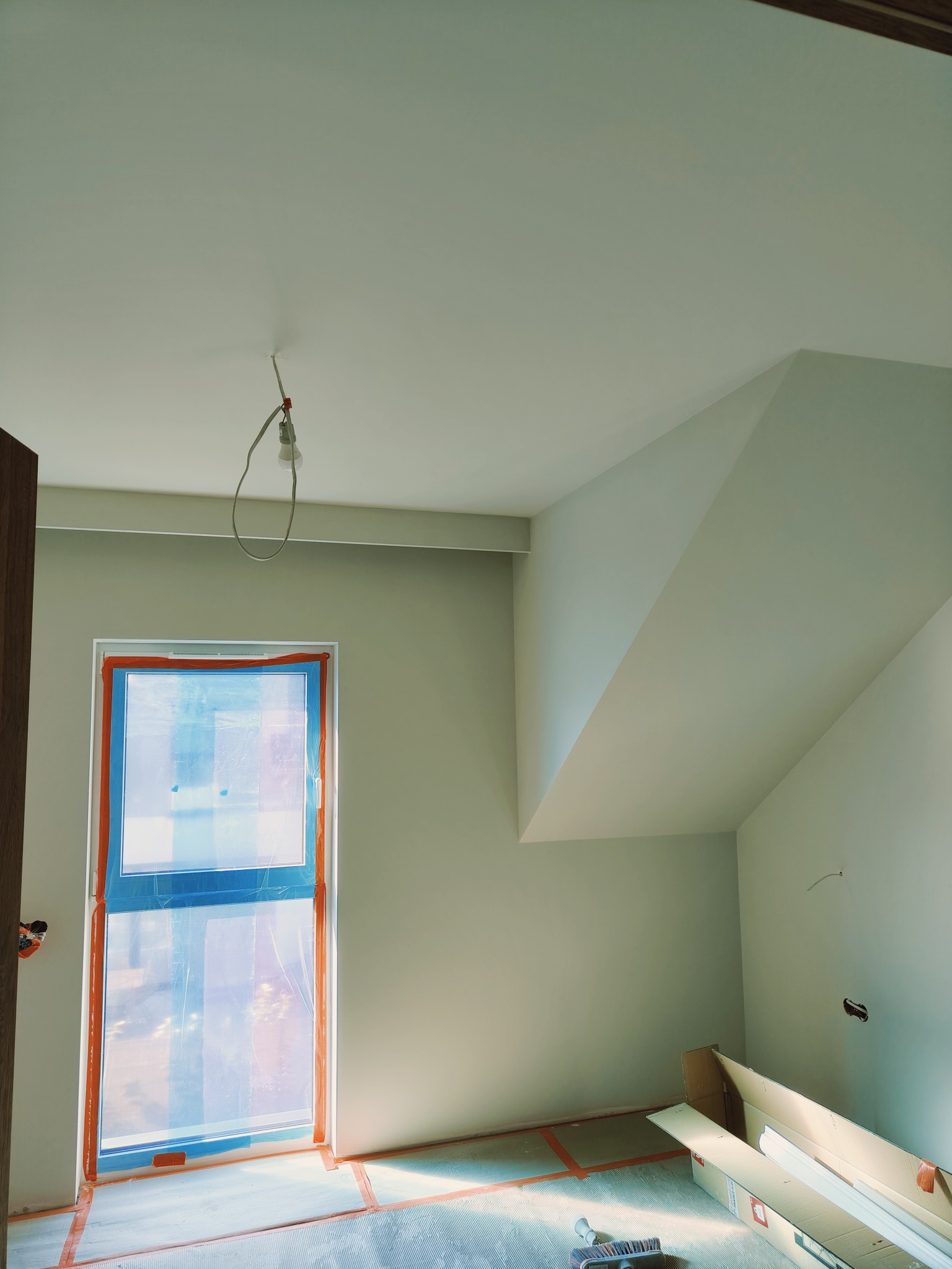 Pokój na poddaszu w trakcie prac wykończeniowych: okno zabezpieczone folią, ściany świeżo pomalowane na jasny odcień, surowe wykończenie pod sufitem, podłoga zabezpieczona kartonem, widoczna...