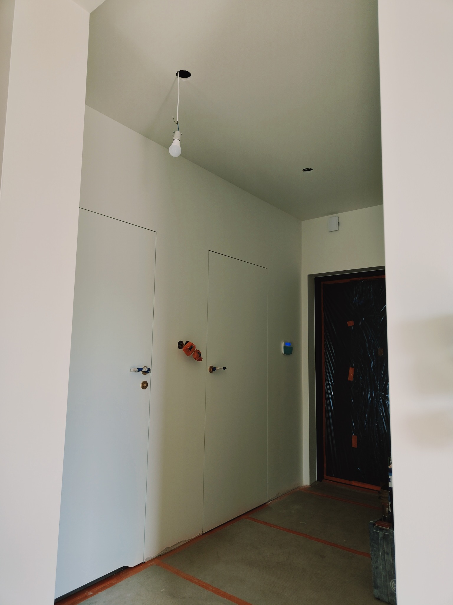 Wnętrze w trakcie prac wykończeniowych: widoczne dwie białe, minimalistyczne drzwi ukryte w ścianie, surowy beton na podłodze z pomarańczowymi liniami wyznaczającymi obszar roboczy, tymczasowa...