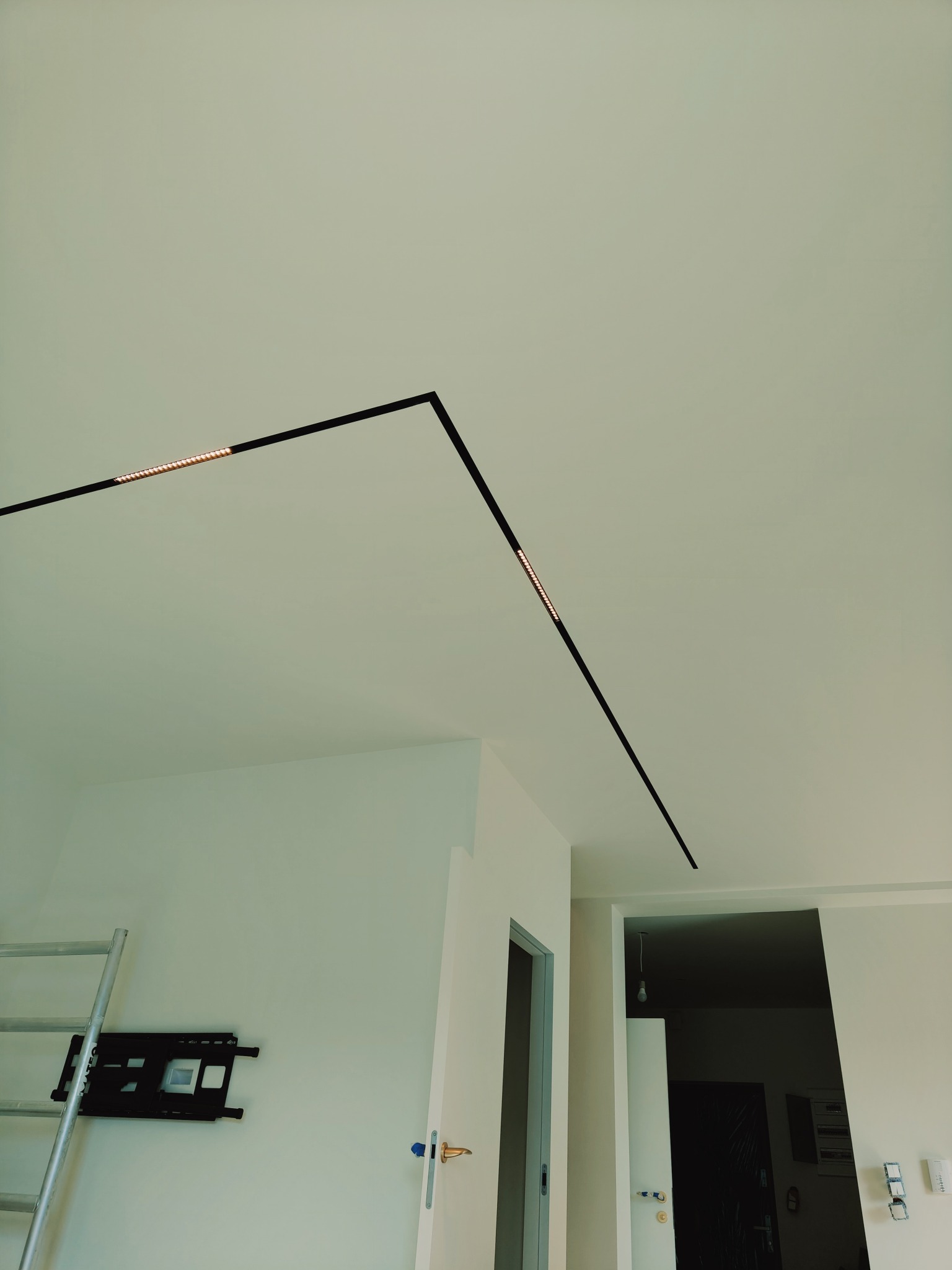 Nowoczesne oświetlenie sufitowe w kształcie litery L, czarna listwa z wbudowanymi punktami świetlnymi, widoczna drabina i uchwyt do telewizora na ścianie.