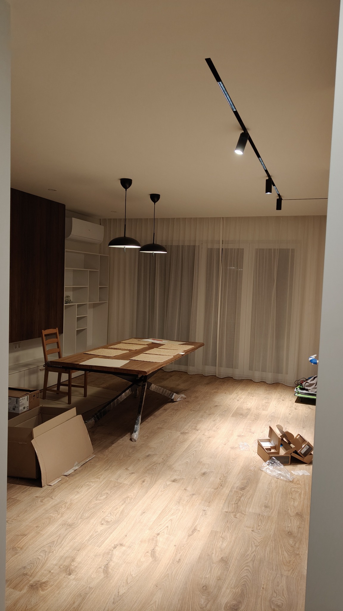 Jasne wnętrze z drewnianą podłogą i stołem. Widoczne oświetlenie sufitowe, firany w oknach i elementy wykończenia.