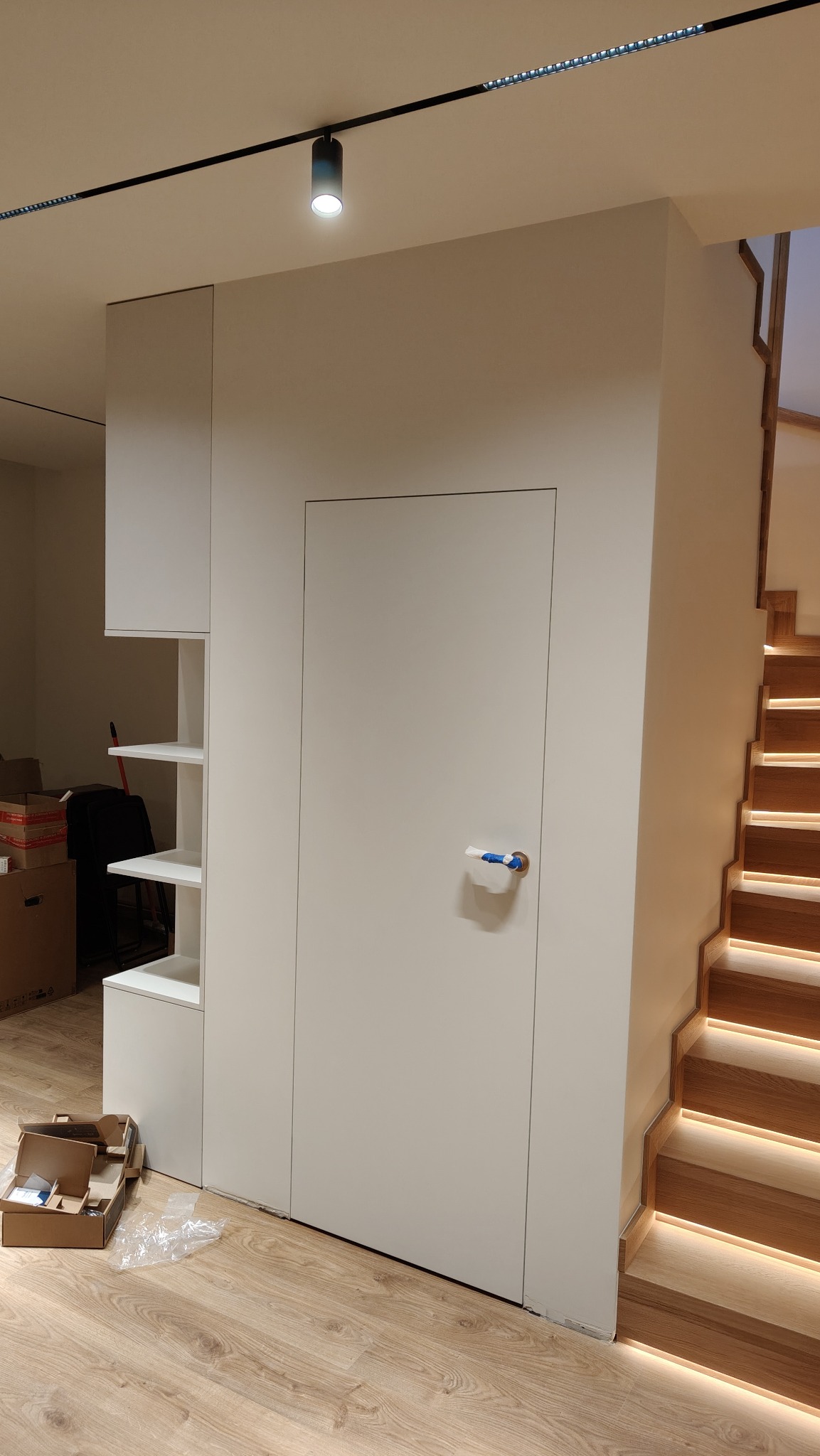 Nowoczesne wykończenie wnętrza: biała zabudowa z ukrytymi drzwiami i oświetlonymi schodami z drewna. Minimalistyczny design, subtelne oświetlenie punktowe.