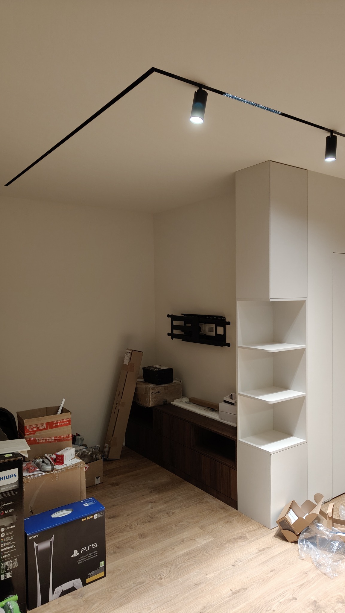 Nowoczesne oświetlenie sufitowe z reflektorami punktowymi na szynie, białe ściany, drewniana podłoga i meble w trakcie montażu, widoczne kartony i elementy wyposażenia.