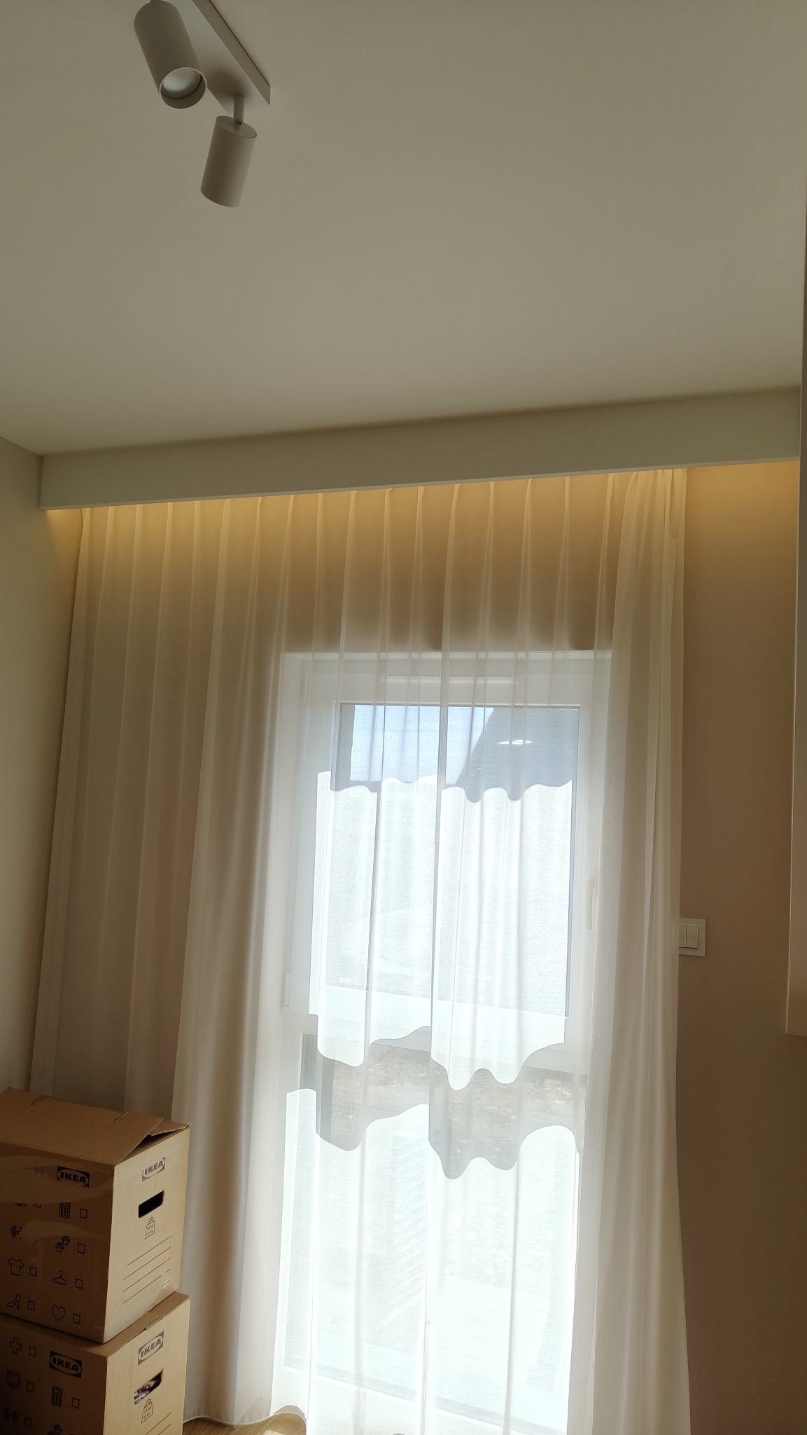 Białe, zwiewne firany z podświetleniem nad oknem w pokoju w Obornikach. Widoczne kartony. Minimalistyczny wystrój wnętrza. Delikatne światło dzienne.