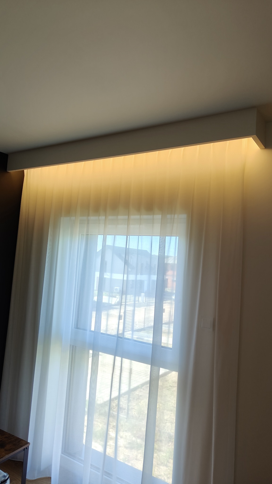 Biała, zwiewna firanka okienna z podświetleniem LED, zamontowana pod sufitem, w nowoczesnym wnętrzu. Widok na zewnątrz przez okno.