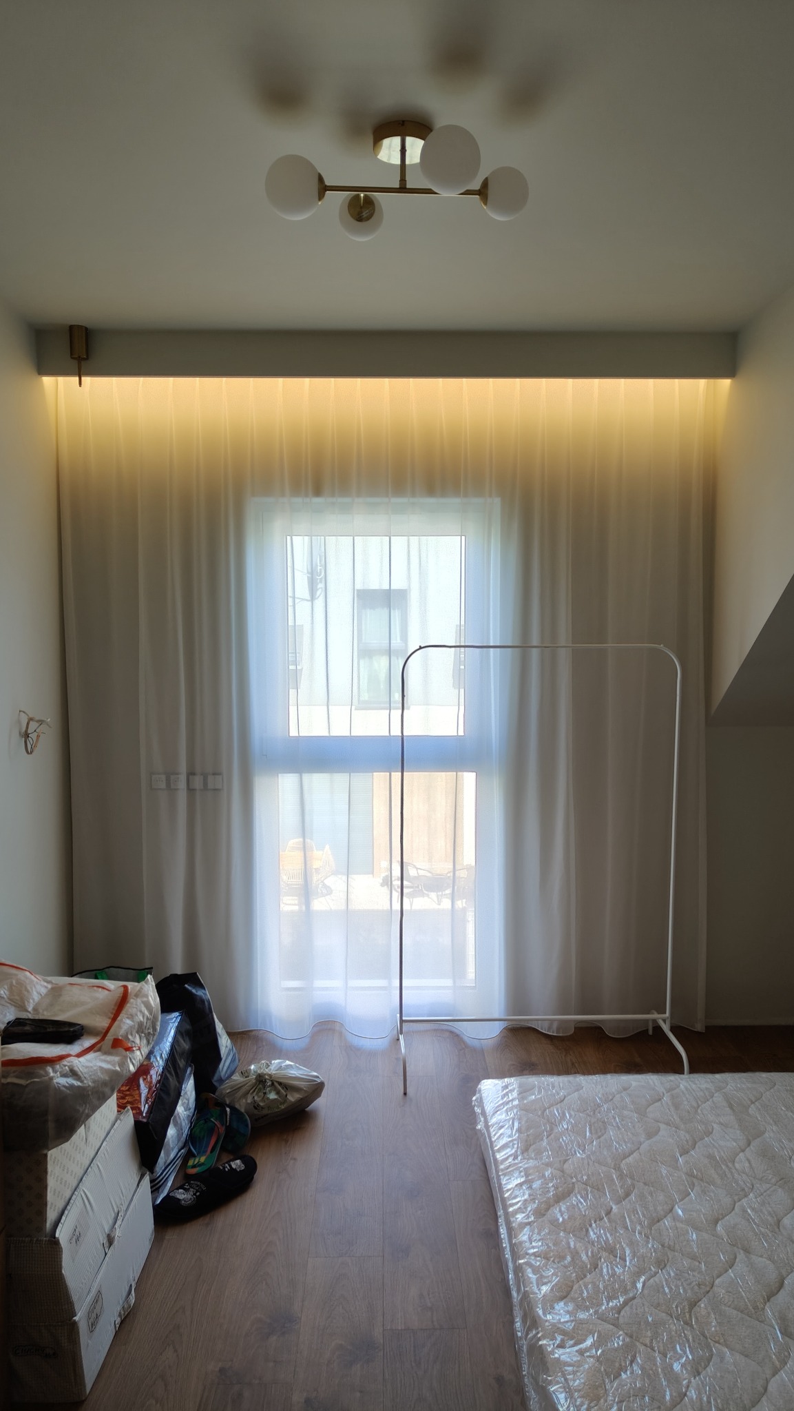 Jasne wnętrze pokoju z oknem zasłoniętym białymi, zwiewnymi zasłonami, oświetlonym paskiem ledowym. Widoczny wieszak na ubrania i spakowane rzeczy.