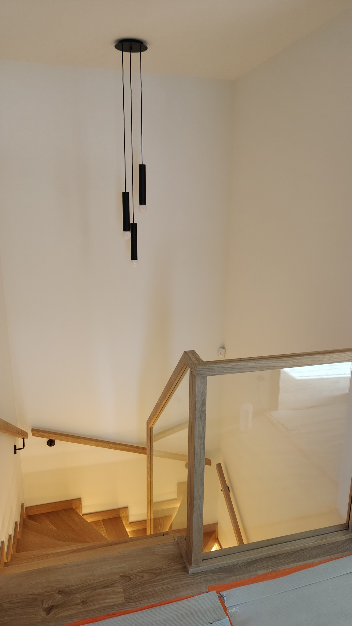 Nowoczesne schody drewniane z szklaną balustradą i minimalistyczną lampą wiszącą. Jasne wnętrze, widok z góry ukazuje detale wykończenia i oświetlenie.