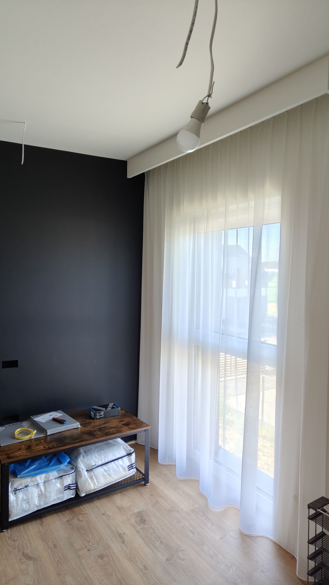 Minimalistyczny salon z czarną ścianą, drewnianą podłogą i białymi, zwiewnymi zasłonami przy oknie. Widoczna lampa wisząca i stolik z akcesoriami.