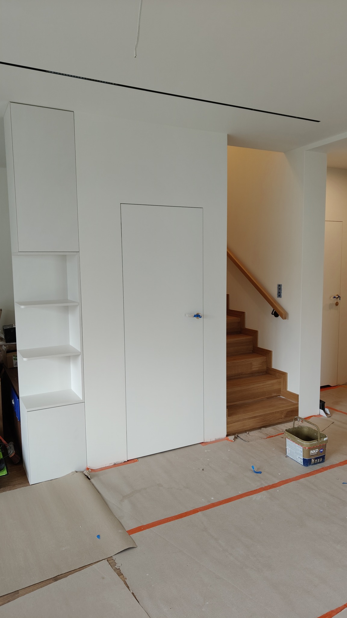 Białe zabudowy meblowe z ukrytymi drzwiami i schodami z drewnianymi stopniami. Podłoga zabezpieczona kartonem i taśmą malarską, widoczne wiadro z farbą.