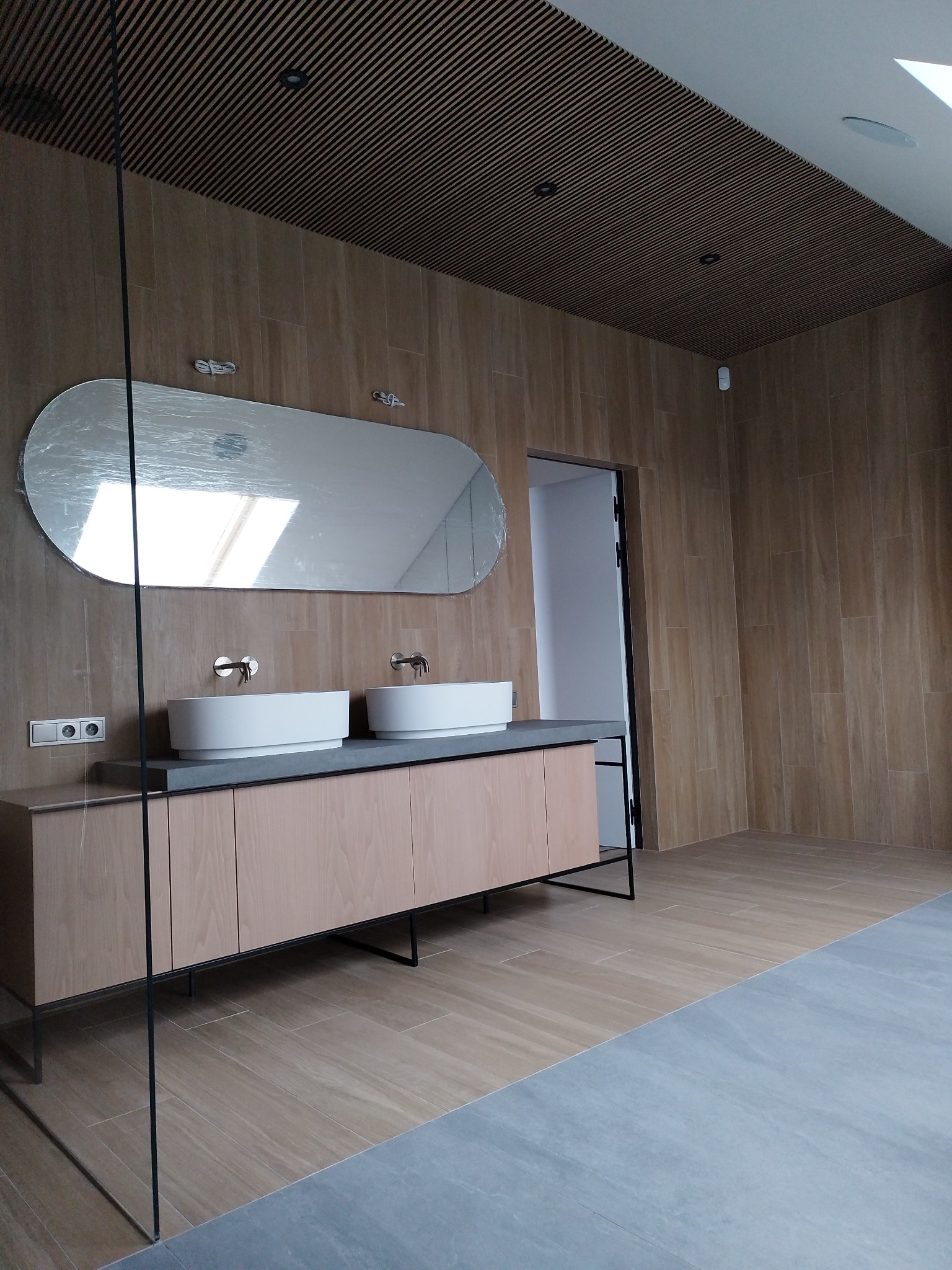 Nowoczesna łazienka z dwoma umywalkami na szafce z drewnianym frontem, owalnym lustrem w folii ochronnej i drewnianym sufitem lamelowym.