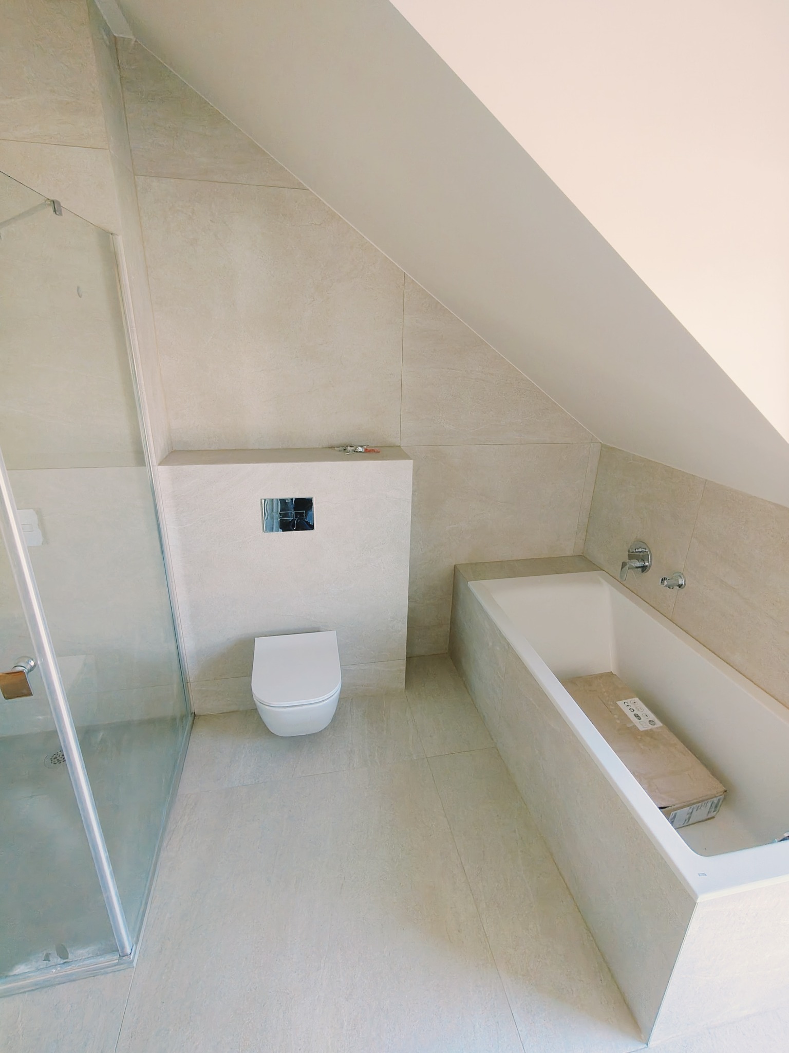 Jasna, wykafelkowana łazienka na poddaszu ze szklaną kabiną prysznicową, podwieszaną toaletą i zabudowaną wanną z armaturą, ukazana z góry.