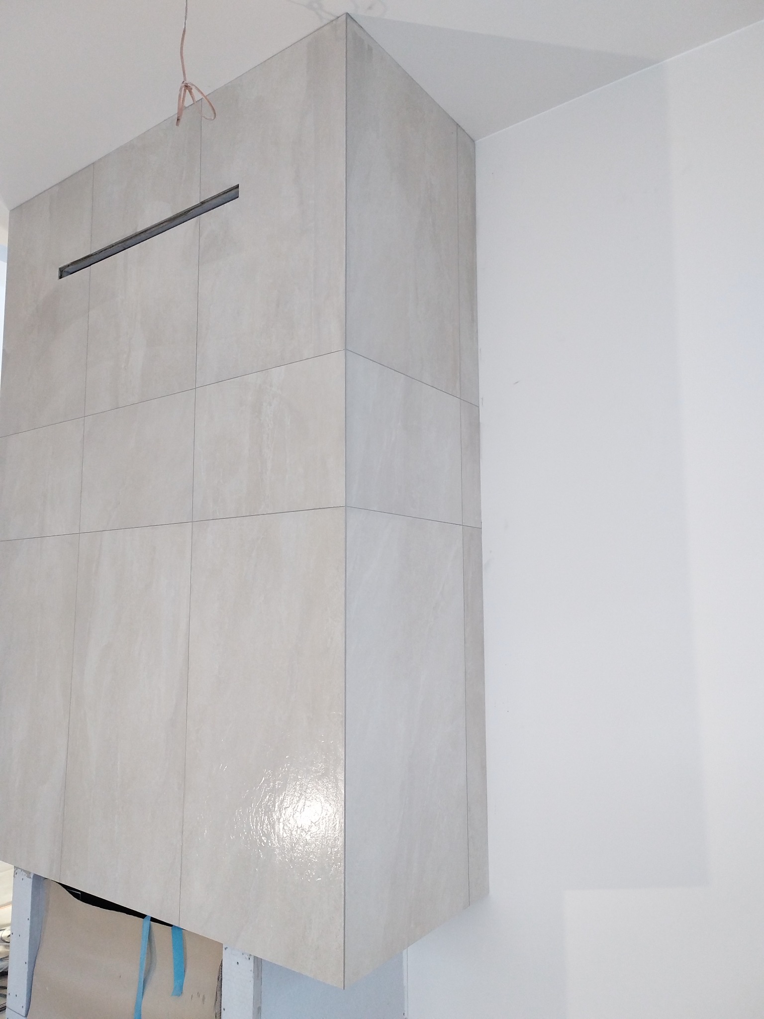 Obudowa kominka z jasnych, kwadratowych płytek, z poziomą szczeliną wentylacyjną, w rogu pomieszczenia. Widoczne niedokończone prace u dołu konstrukcji.