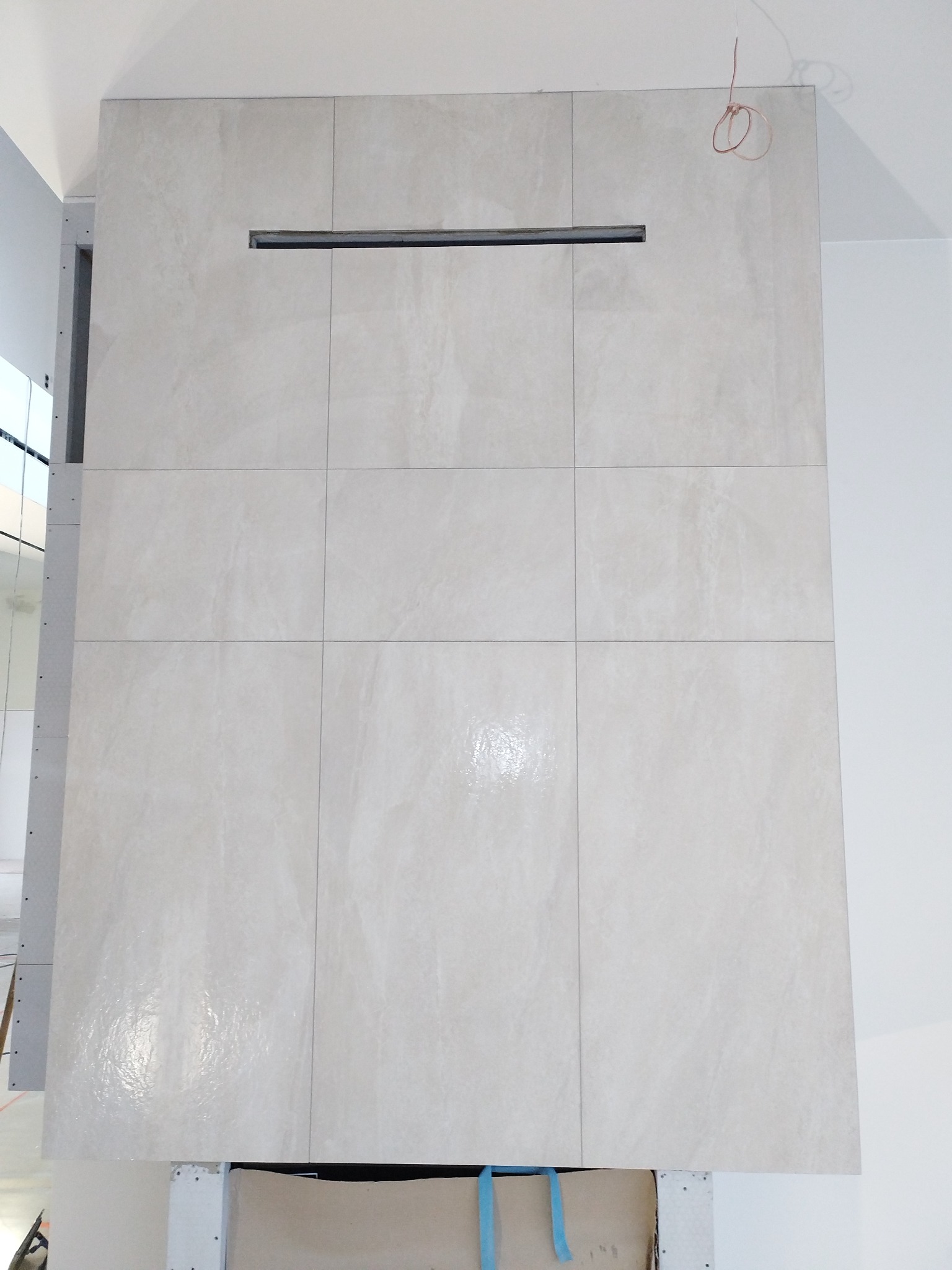 Wykończona ściana z jasnych, kwadratowych płytek ceramicznych, z poziomym otworem wentylacyjnym. Widoczne fragmenty sufitu i ściany bocznej.