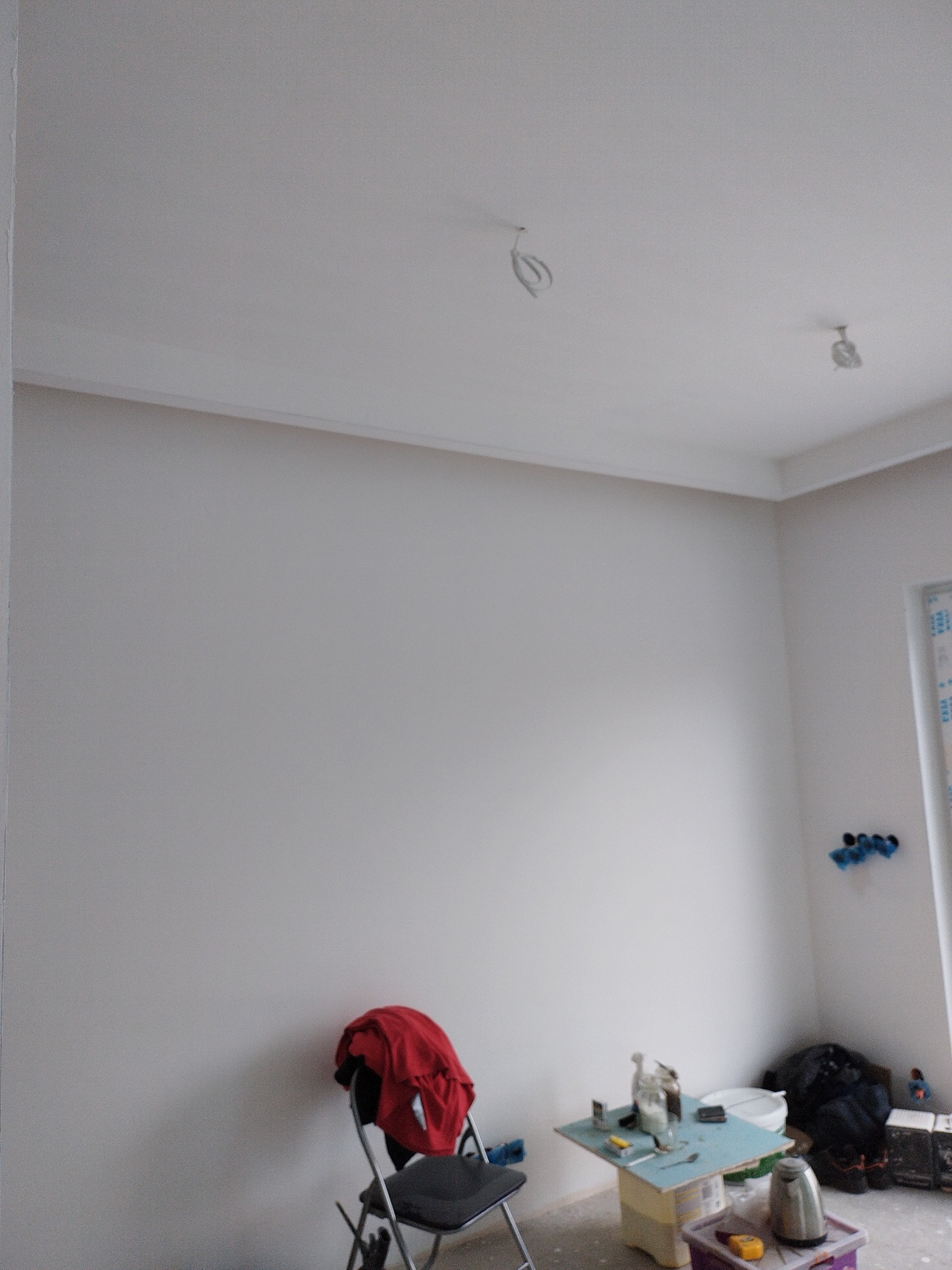 Świeżo pomalowane, jasnoszare ściany i sufit w pokoju. Widoczne kable wystające z sufitu, krzesło z czerwoną kurtką i stolik z narzędziami w rogu pomieszczenia.