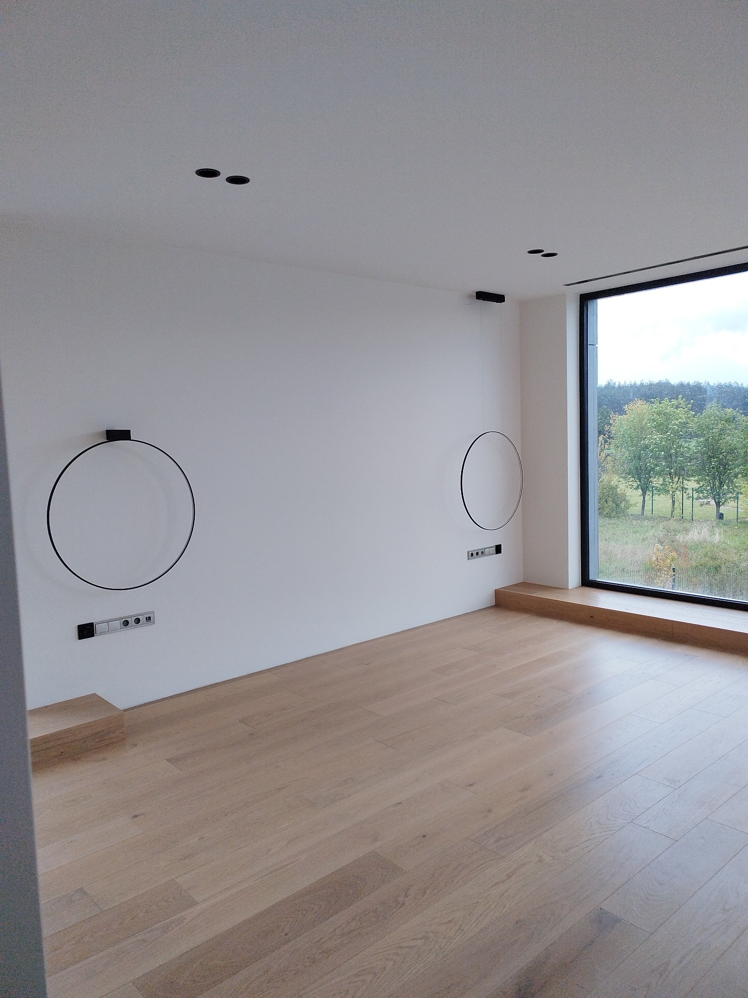 Minimalistyczne wnętrze z drewnianą podłogą, białymi ścianami i dużym oknem z widokiem na zieleń. Dekoracyjne, okrągłe lampy ścienne.