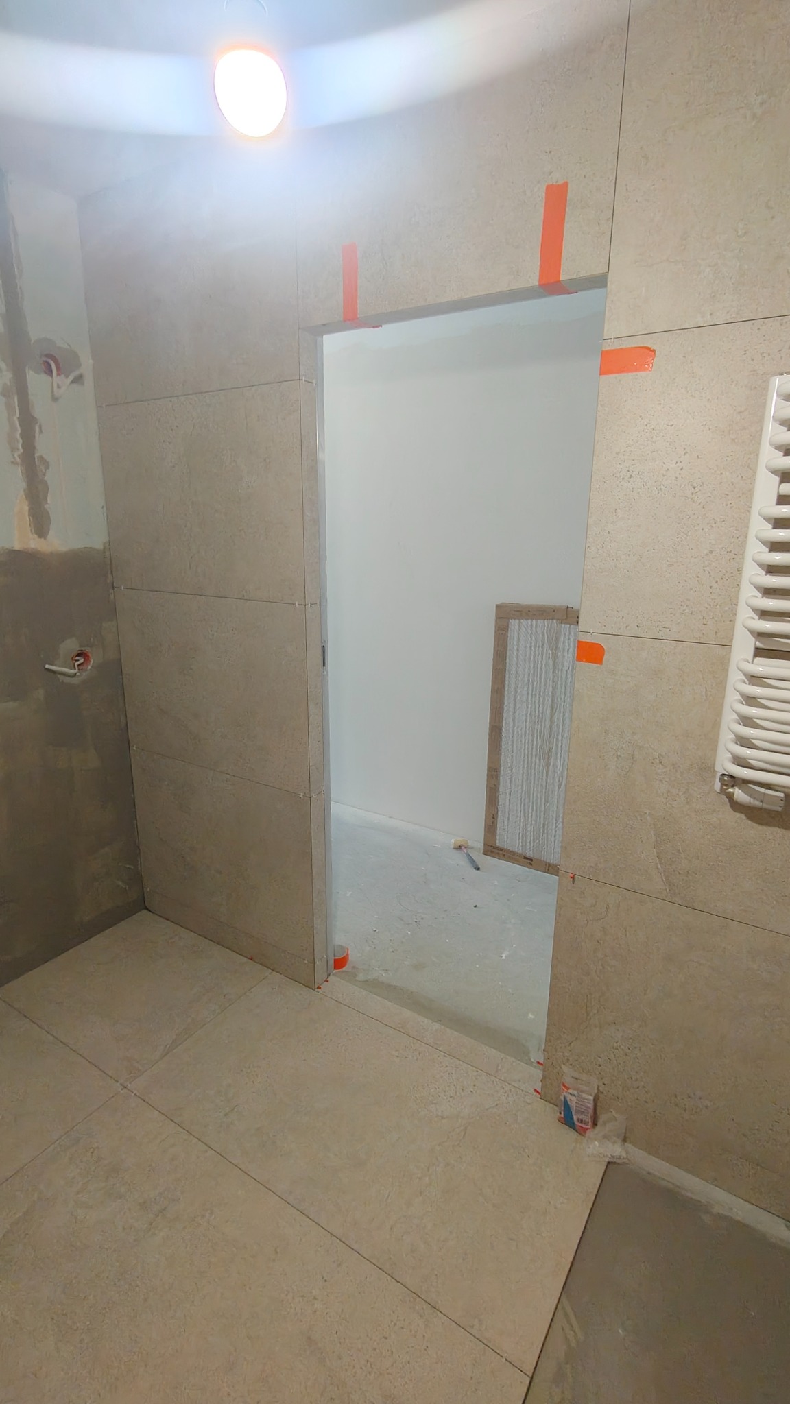 Układanie płytek w łazience: beżowe płytki na ścianach i podłodze, widoczne narzędzia i fragment grzejnika. Prace w toku, z użyciem taśmy maskującej.