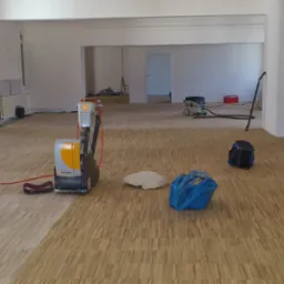 Profesjonalna maszyna Pallmann szlifuje parkiet w przestronnym, jasnym pomieszczeniu; widoczna różnica w kolorze między obszarem obrobionym a nieobrobionym; w tle odkurzacz przemysłowy i torba...