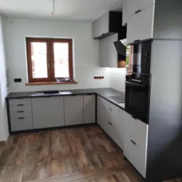 Nowoczesna kuchnia w odcieniach szarości i czerni z drewnianą podłogą, widoczne okno z drewnianą ramą i fragment instalacji elektrycznej na suficie.