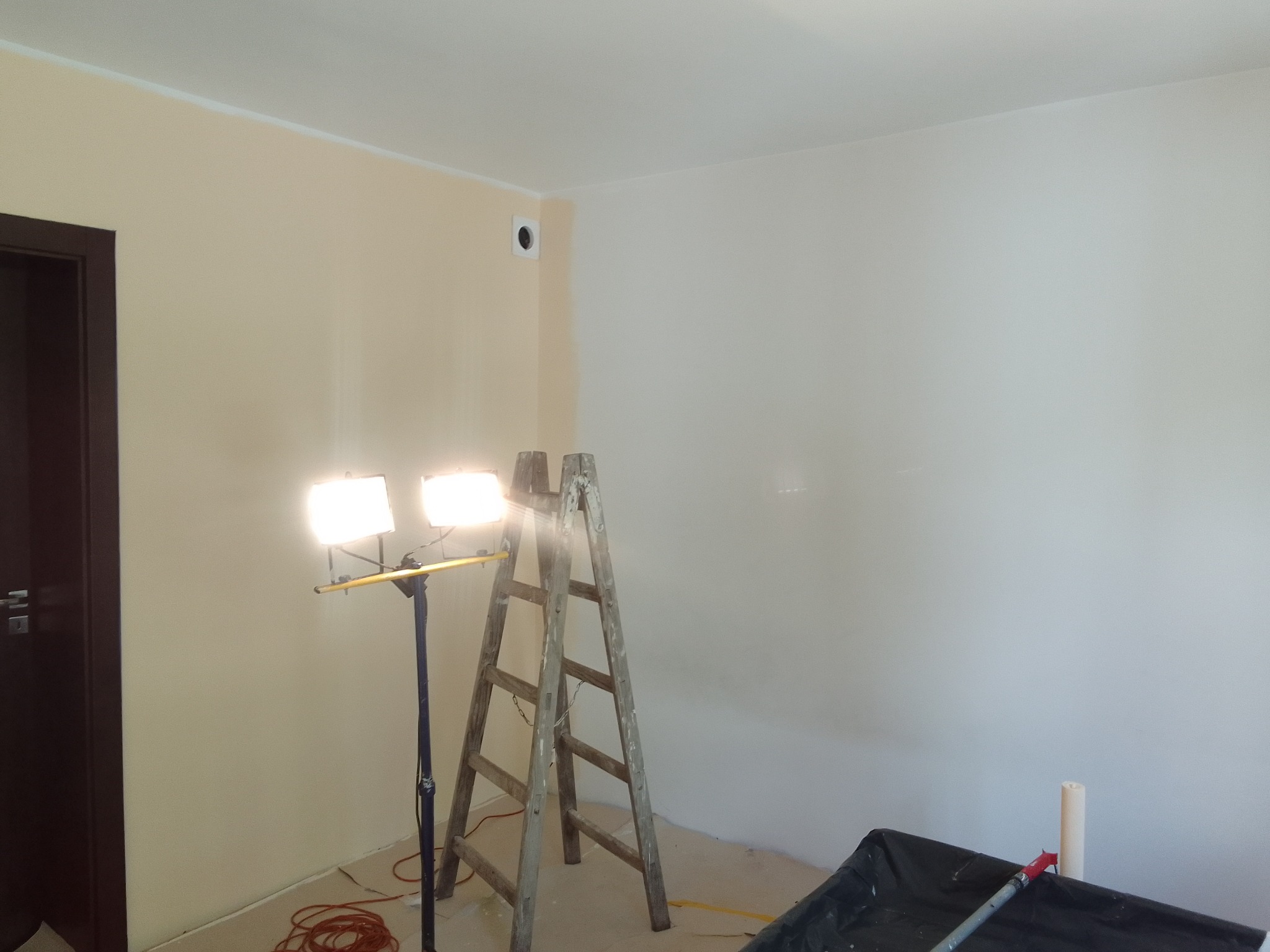 Pokój w trakcie odświeżania, ściany częściowo pomalowane na beżowo i biało, widoczna drabina, lampa halogenowa na statywie i zabezpieczenia podłogi.