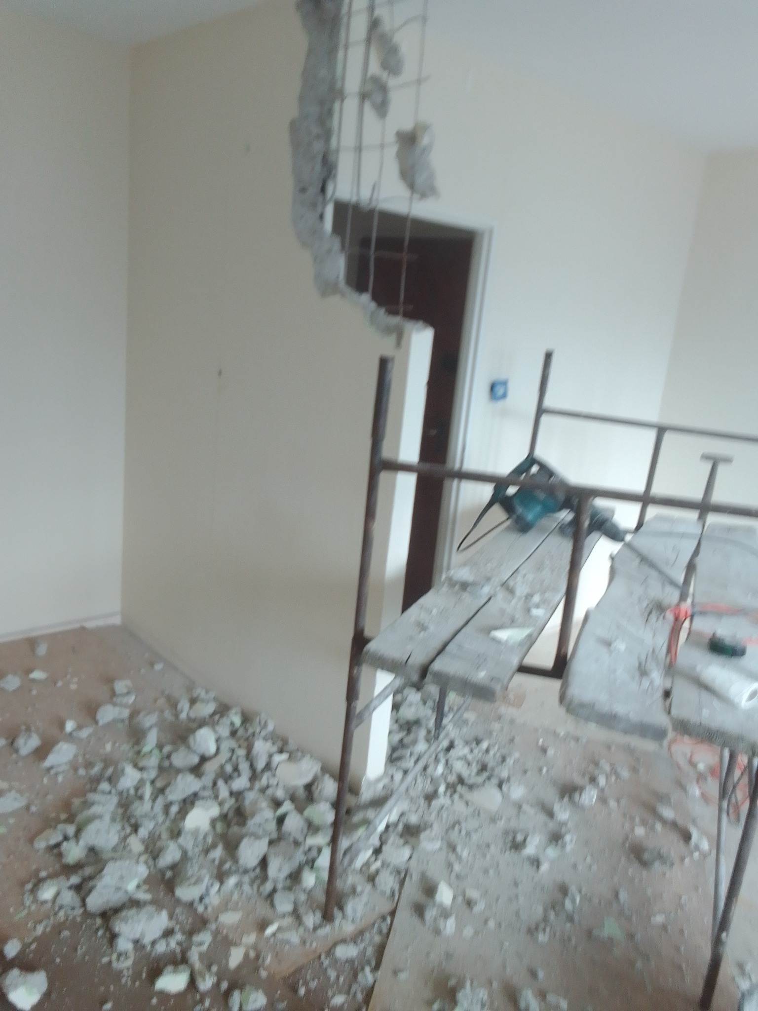 Fragment ściany z odsłoniętym zbrojeniem, gruz na podłodze, rusztowanie z wiertarką, remont w trakcie.