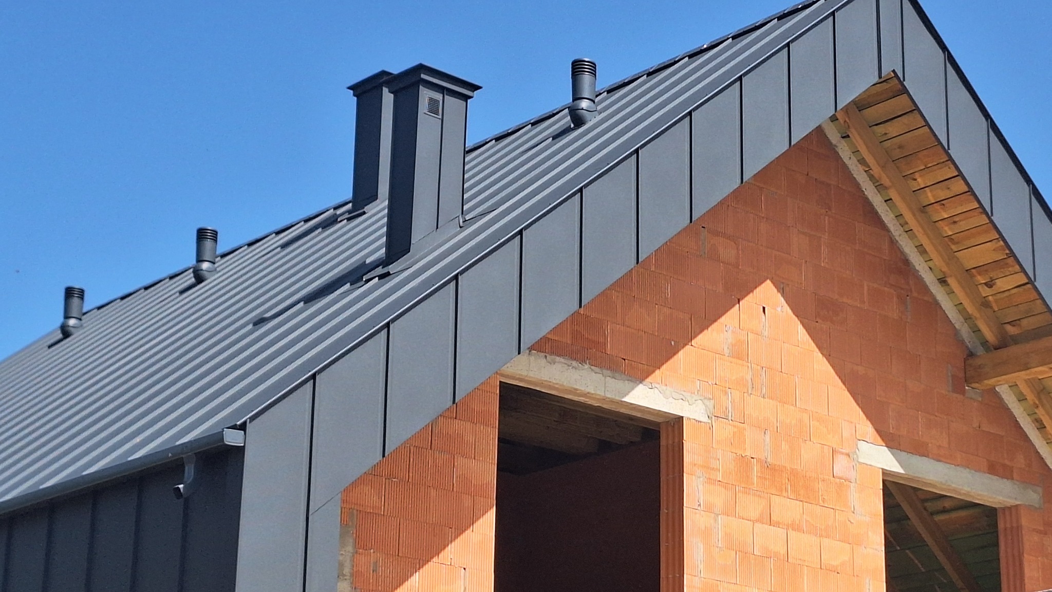 Szary dach z blachy na budynku w trakcie budowy, widoczne kominy i fragment ściany z cegły dziurawki, błękitne niebo w tle.