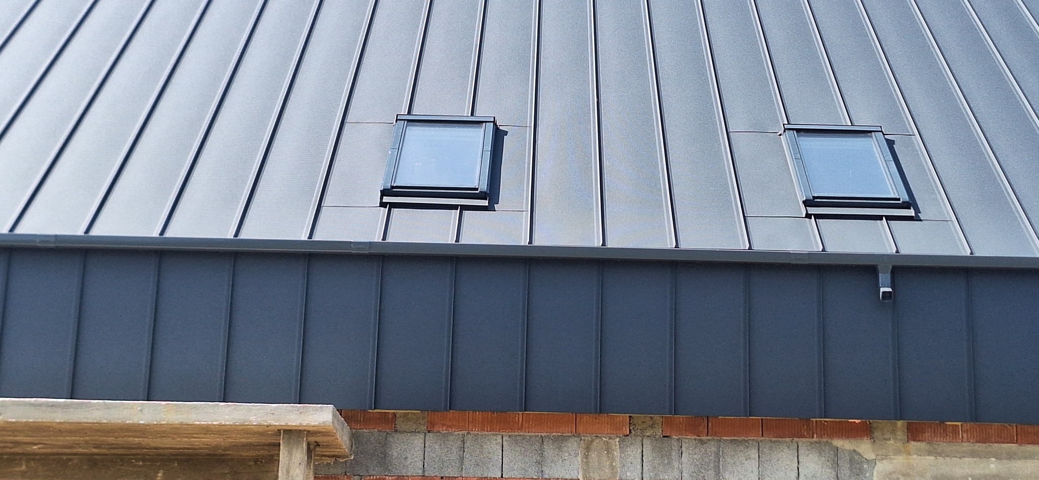 Szary dach z blachy na rąbek stojący z dwoma oknami dachowymi. Widoczne fragmenty muru i rusztowania.