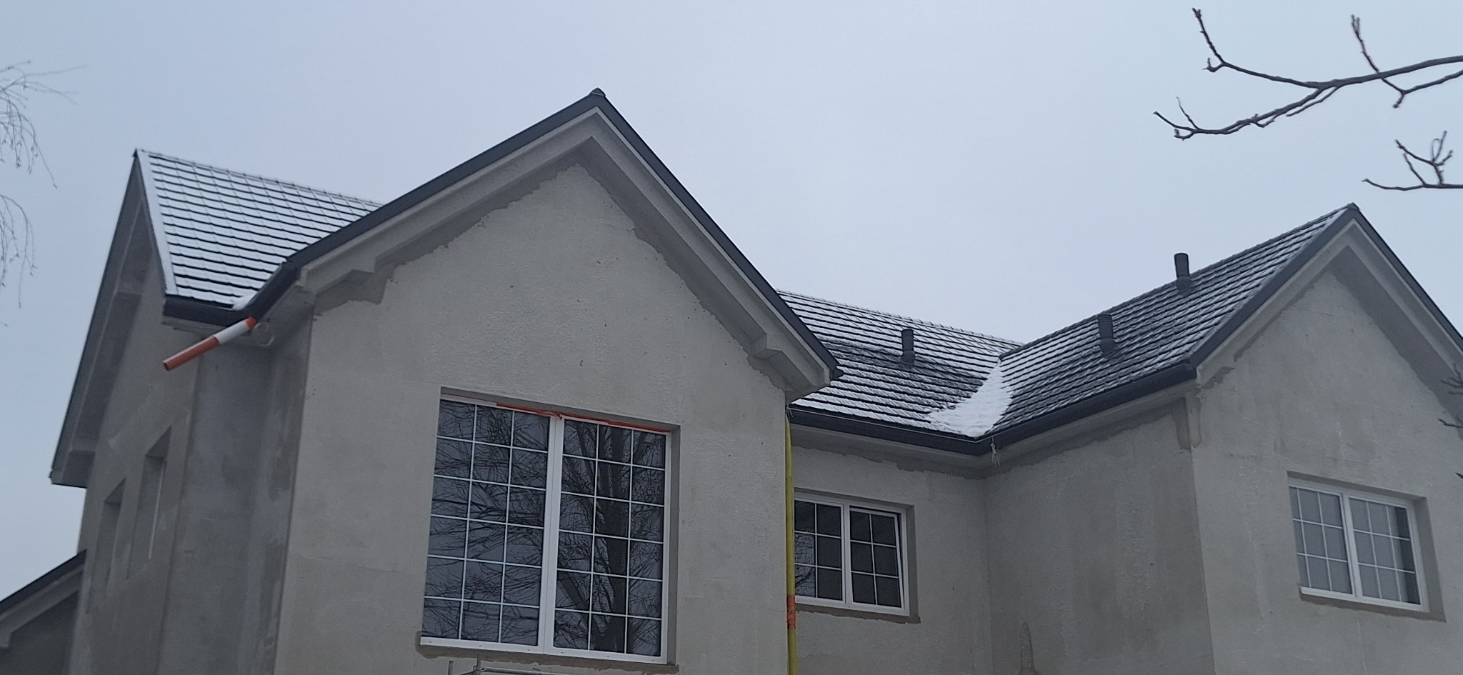 Dom w trakcie wykańczania z ciemnym dachem pokrytym śniegiem. Widoczne rynny i okna z białymi ramami. Szary, pochmurny dzień.