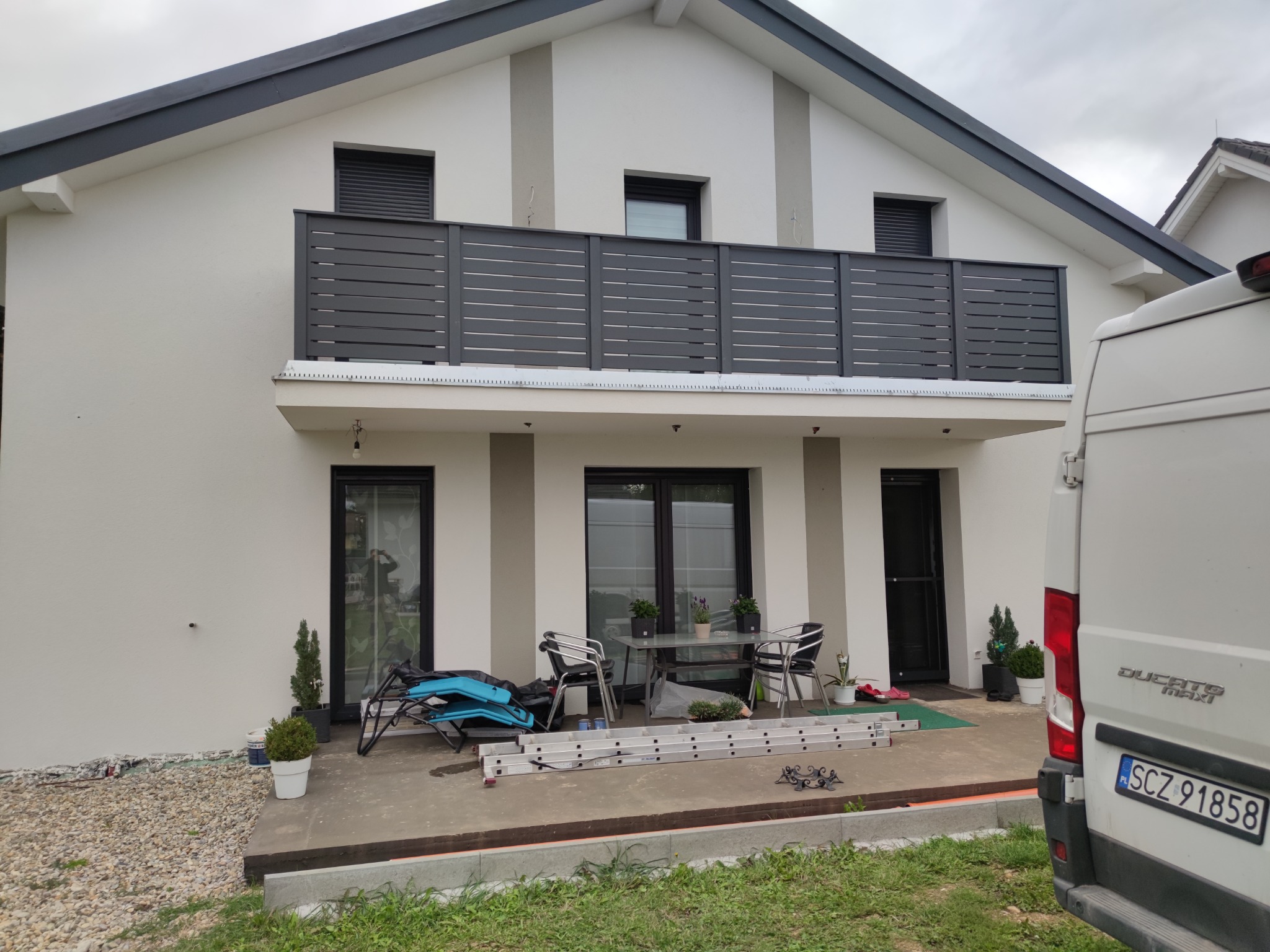Biały dom z szarą, poziomą balustradą na balkonie. Na tarasie widoczne meble ogrodowe, drabina i częściowo zaparkowany biały samochód dostawczy.