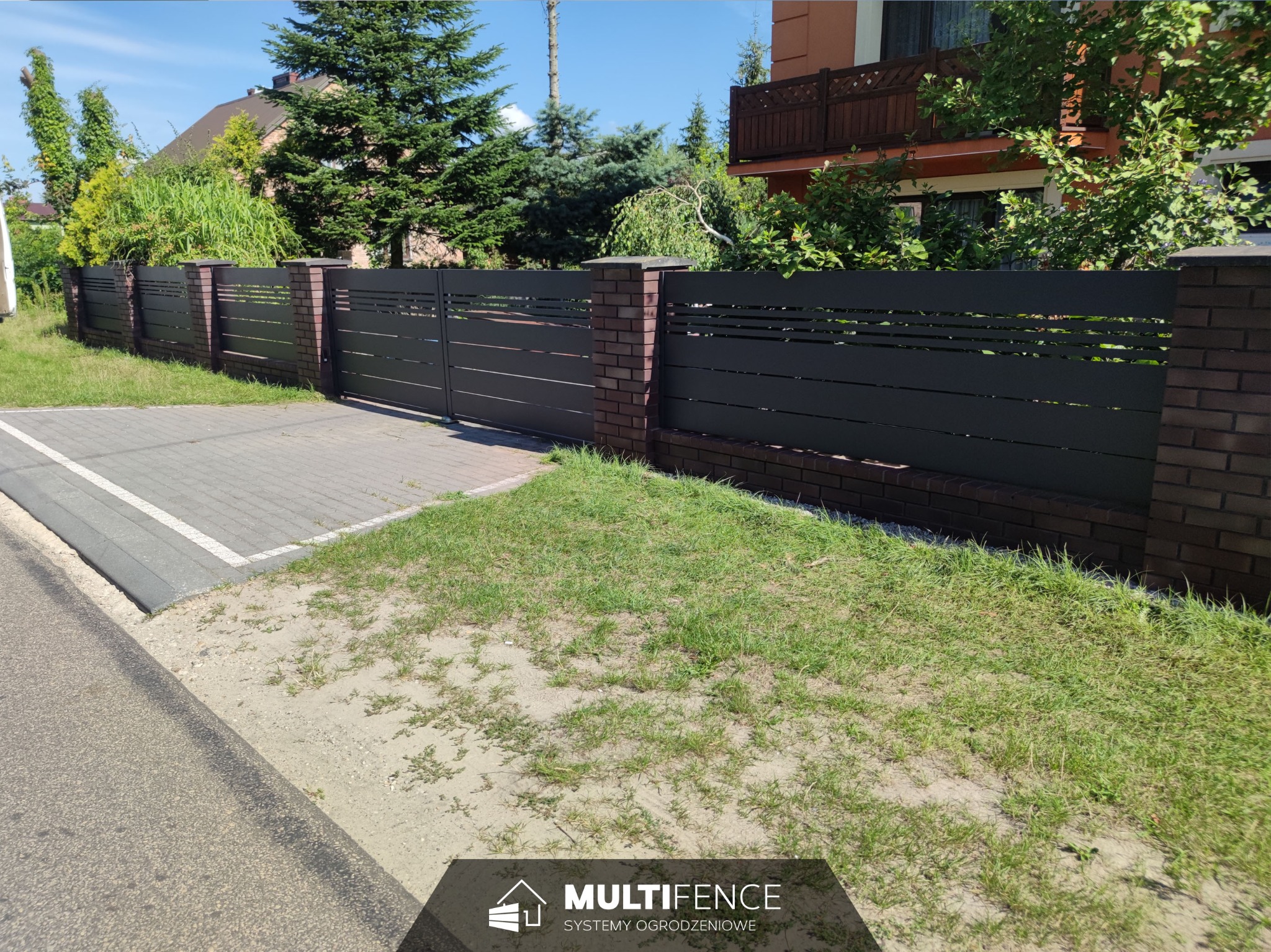 Nowoczesne ogrodzenie z poziomych, szarych paneli z prześwitami, osadzone na ceglanych słupkach, z bramą wjazdową, na tle zieleni i domu.