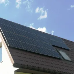 Panele słoneczne zamontowane na brązowym dachu domu jednorodzinnego z oknem dachowym, widoczne białe ściany i błękitne niebo z chmurami.