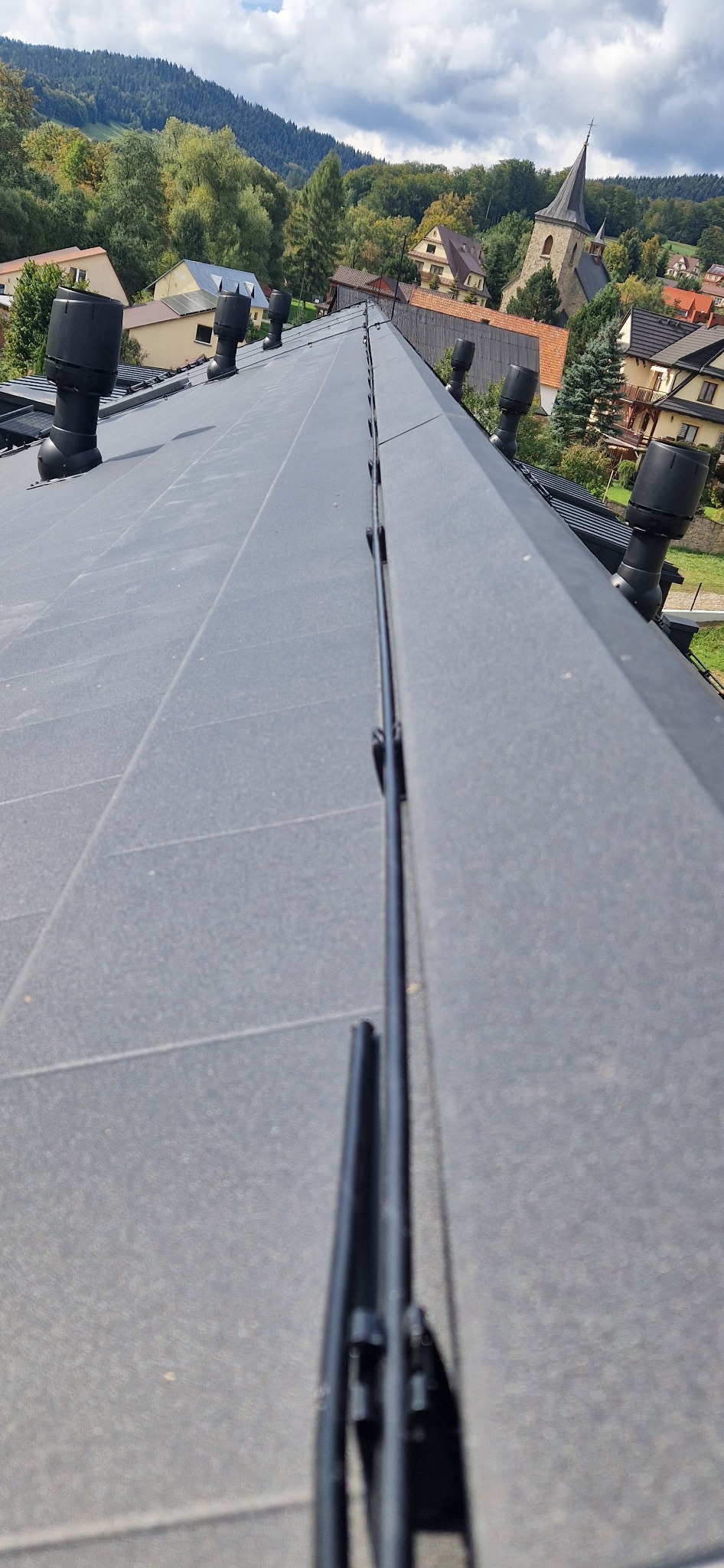 Szary dach z ceramicznej dachówki z zamontowaną instalacją odgromową w postaci czarnego przewodu z uchwytami, kominki wentylacyjne na dachu, w tle zabudowa mieszkalna i zalesione wzgórza.