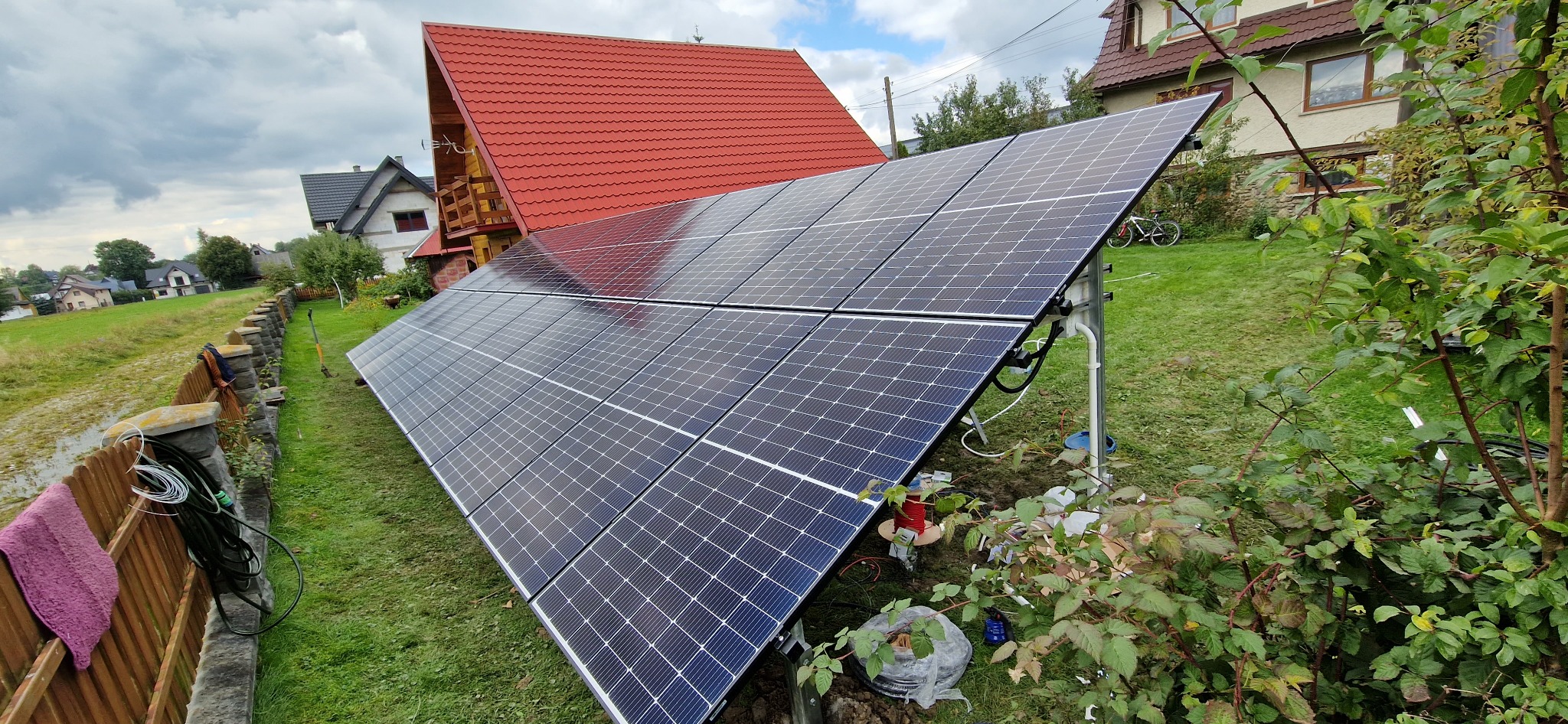 Panele fotowoltaiczne zamontowane w ogrodzie, widoczne kable, konstrukcja wsporcza i elementy montażowe, w tle dom z czerwoną dachówką.