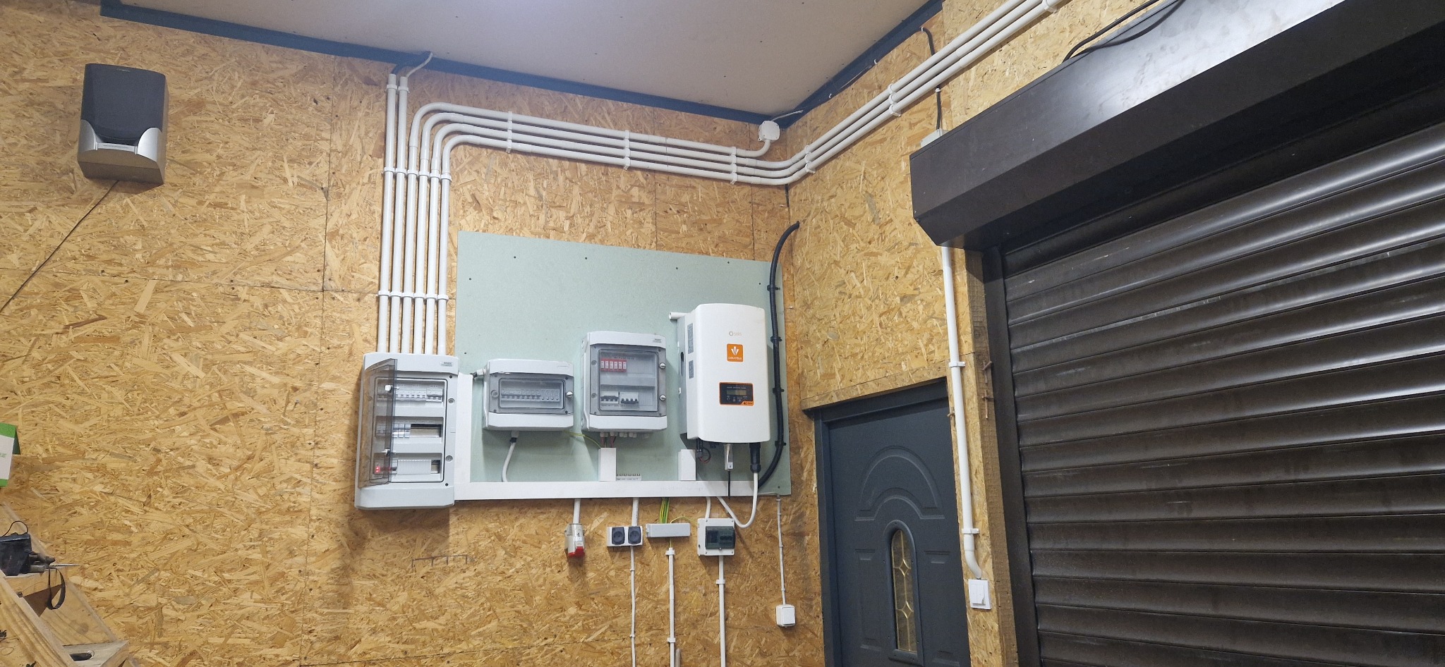 Rozdzielnice elektryczne, inwerter i kable w białych rurkach na płycie OSB w pomieszczeniu z roletą antywłamaniową.