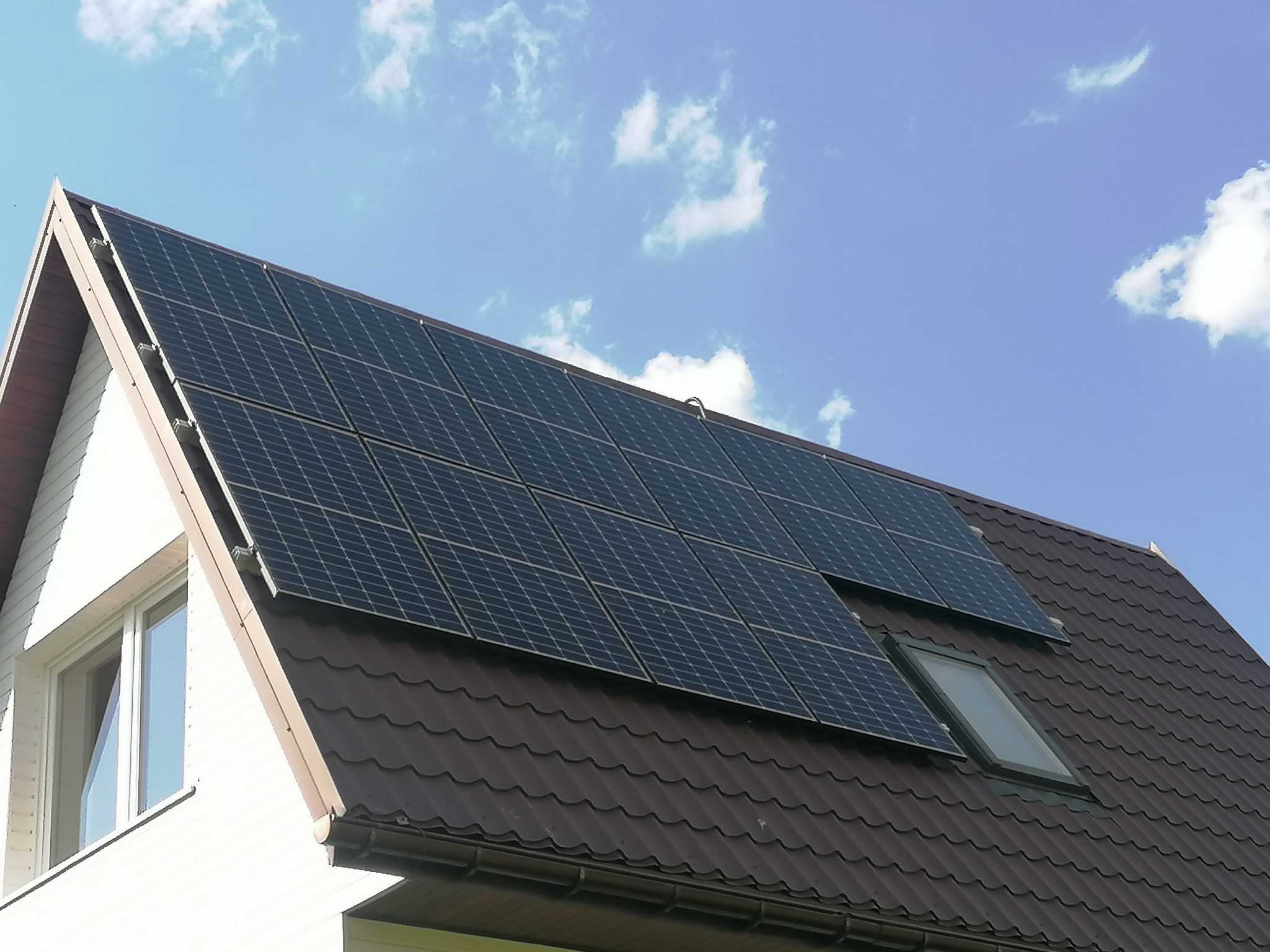 Panele słoneczne zamontowane na brązowym dachu domu jednorodzinnego z oknem dachowym, widoczne białe ściany i błękitne niebo z chmurami.