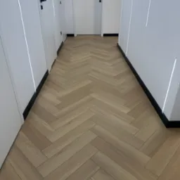 Jasny parkiet ułożony w jodełkę w nowoczesnym korytarzu z białymi ścianami i drzwiami z czarnymi klamkami oraz subtelnym oświetleniem LED przy podłodze i na ścianach.