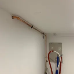 Miedziana rura grzewcza przymocowana do ściany z widocznymi elementami montażowymi, w tle rury PEX w kolorach czerwonym, białym i niebieskim, oraz otwór w ścianie z widoczną płytą gipsową.