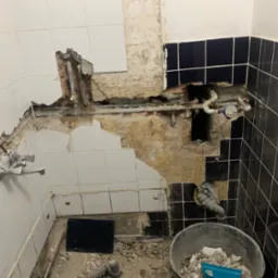Uszkodzona ściana w łazience z widocznymi rurami, fragmentami płytek i gruzem, przygotowana do naprawy instalacji hydraulicznej.