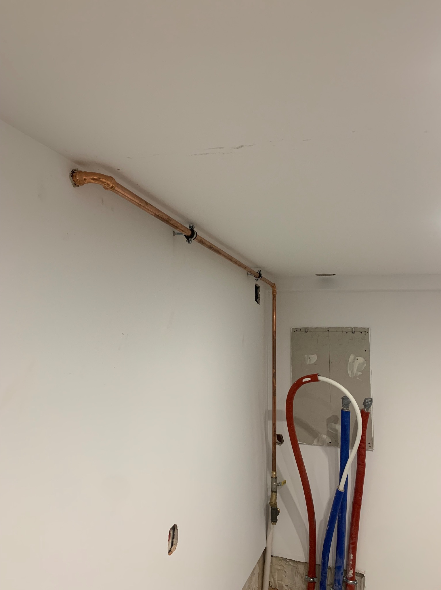 Miedziana rura grzewcza przymocowana do ściany z widocznymi elementami montażowymi, w tle rury PEX w kolorach czerwonym, białym i niebieskim, oraz otwór w ścianie z widoczną płytą gipsową.