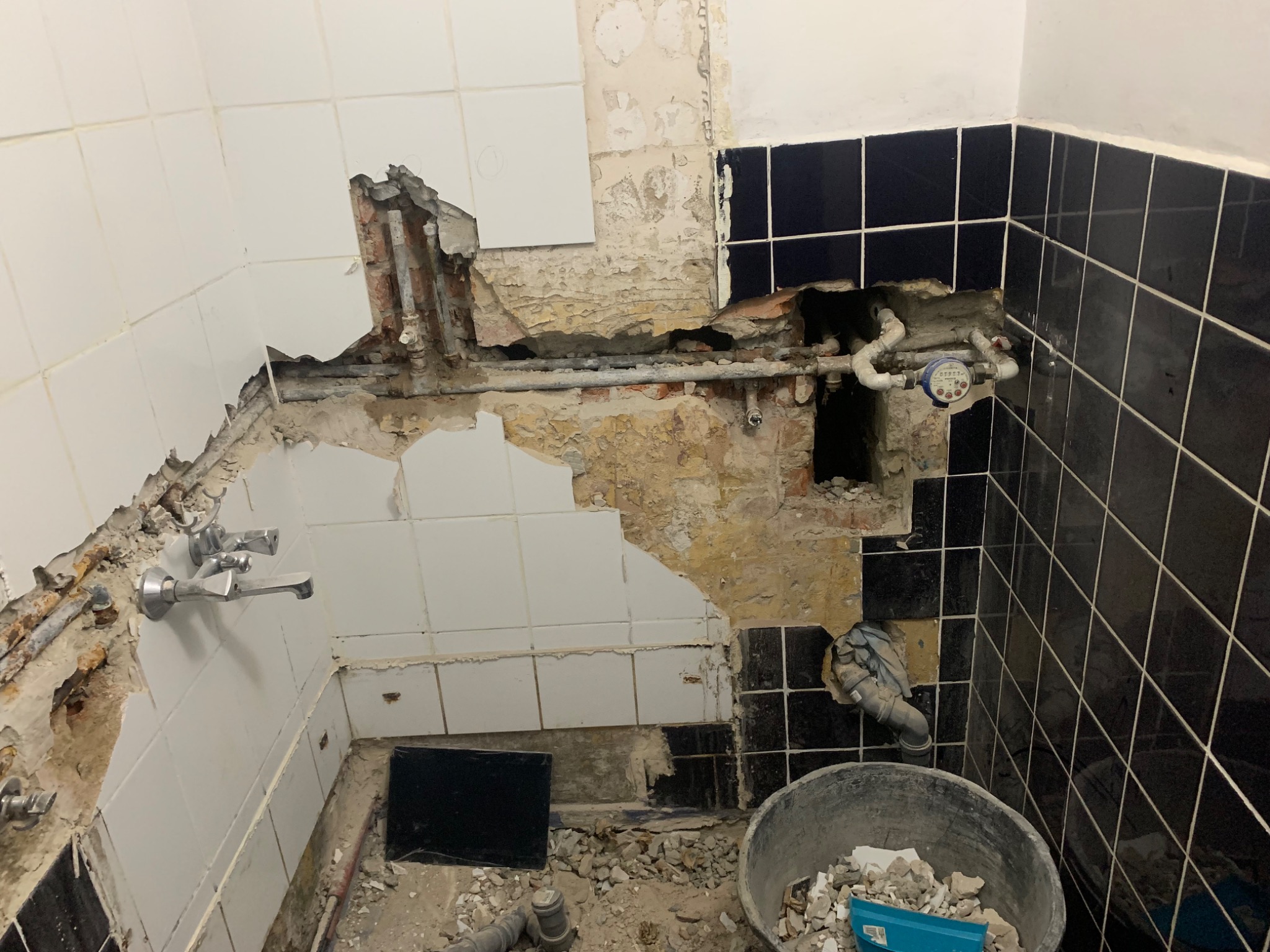 Uszkodzona ściana w łazience z widocznymi rurami, fragmentami płytek i gruzem, przygotowana do naprawy instalacji hydraulicznej.