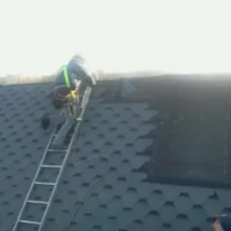 Dekarz w Świnoujściu zabezpieczony uprzężą, w trakcie wchodzenia po drabinie na dach pokryty nową, ciemną dachówką bitumiczną w kształcie plastra miodu. Część dachu jeszcze niepokryta.