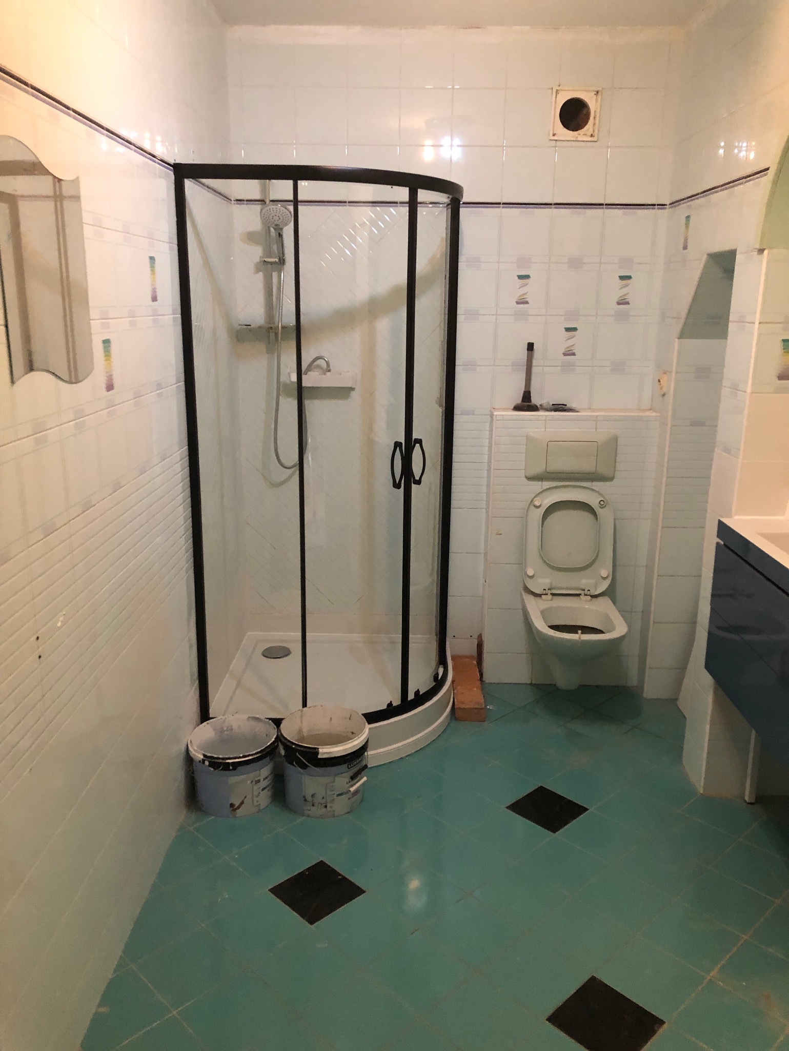 Łazienka z kabiną prysznicową z czarnymi ramami, toaleta z otwartą klapą i wiaderkami po farbie, sugerująca remont w trakcie.