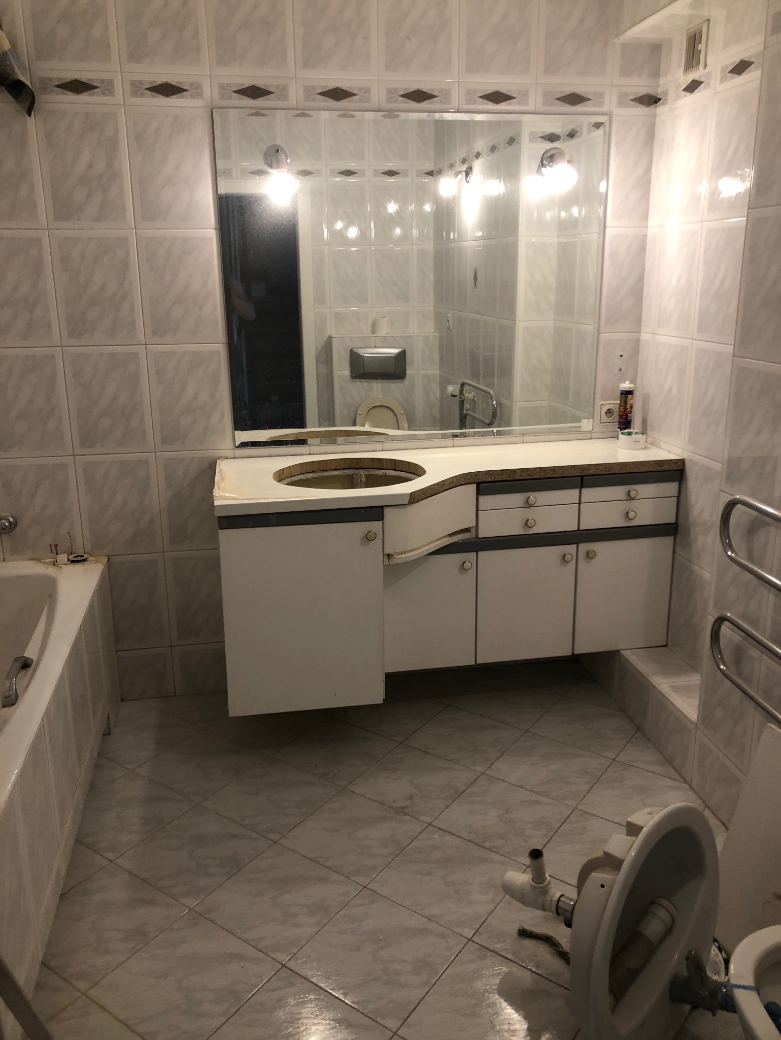 Łazienka w trakcie remontu w Warszawie: zdemontowana umywalka z szafką, widoczne płytki ceramiczne na ścianach i podłodze, fragment wanny oraz zdemontowana toaleta na podłodze.