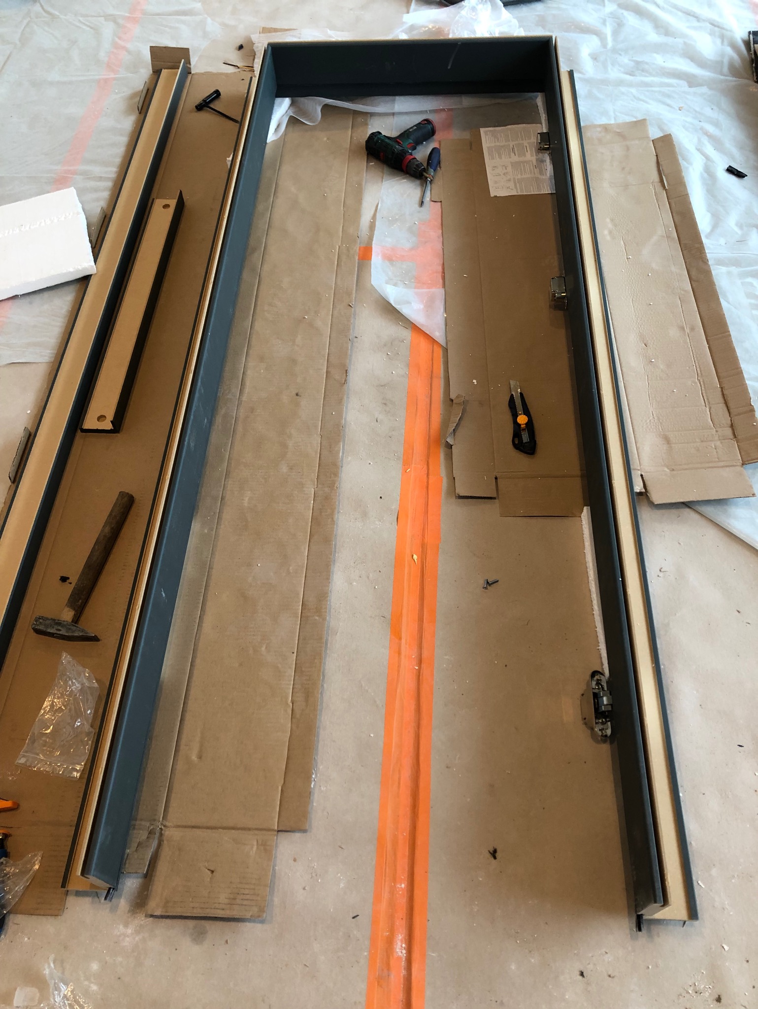 Przygotowane ościeżnice drzwiowe w trakcie montażu, widoczne narzędzia: wkrętarka, nóż tapicerski, młotek, klucz imbusowy, śruby, kartony zabezpieczające podłogę.