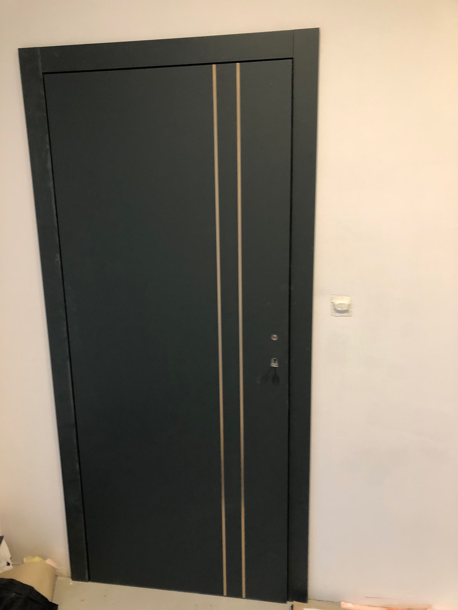 Ciemnoszare drzwi wewnętrzne z dwoma pionowymi, jasnymi paskami, widoczne zamek i klamka, biała ściana w tle.