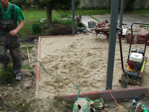 Przygotowanie podłoża: ubity piasek w obramowaniu z obrzeży, zagęszczarka wibracyjna, częściowo widoczny pracownik z grabiami w tle.