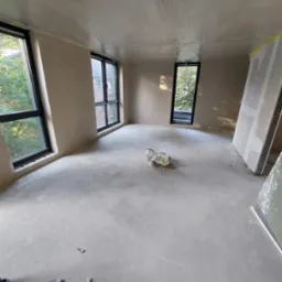 Wnętrze w trakcie prac wykończeniowych: świeżo położony tynk na ścianach i suficie zabezpieczonym folią, widoczne okna z czarnymi ramami, worek zaprawy na podłodze i elementy instalacji elektrycznej.