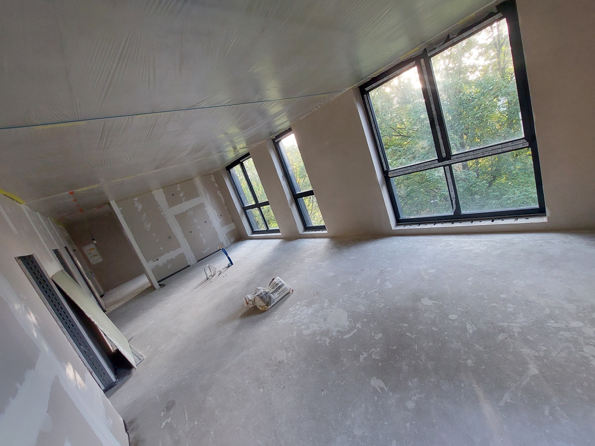 Wnętrze w stanie surowym po wylewce, z widocznymi oknami, zabezpieczonym folią sufitem i workiem zaprawy na podłodze.