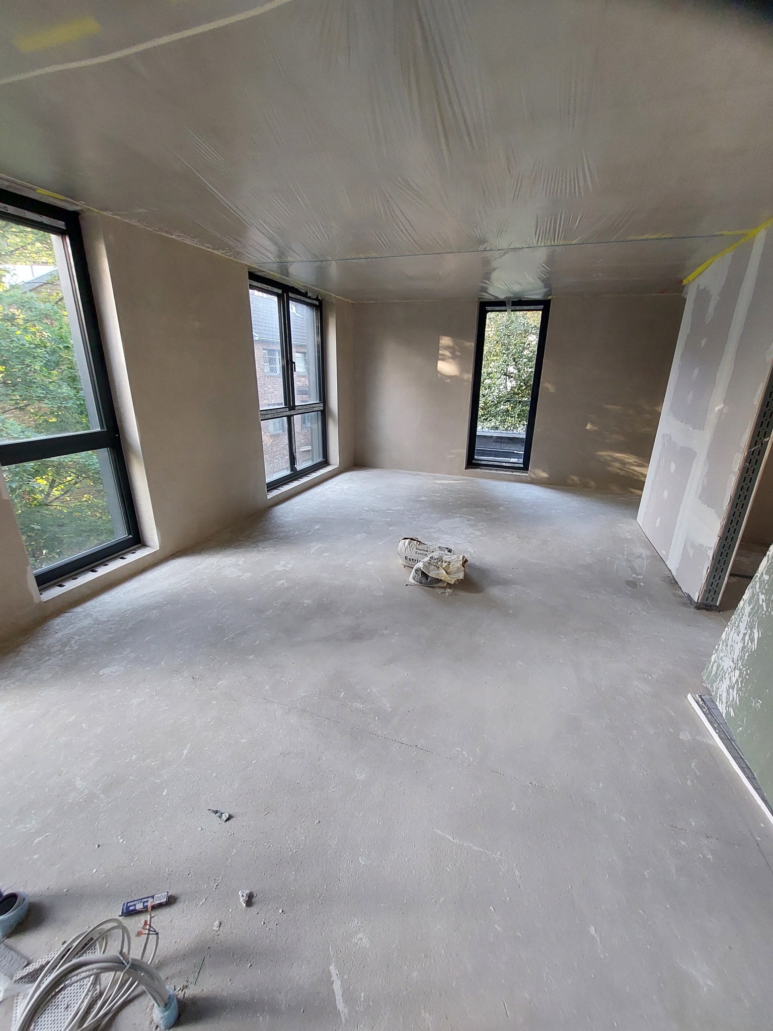 Wnętrze w trakcie prac wykończeniowych: świeżo położony tynk na ścianach i suficie zabezpieczonym folią, widoczne okna z czarnymi ramami, worek zaprawy na podłodze i elementy instalacji elektrycznej.