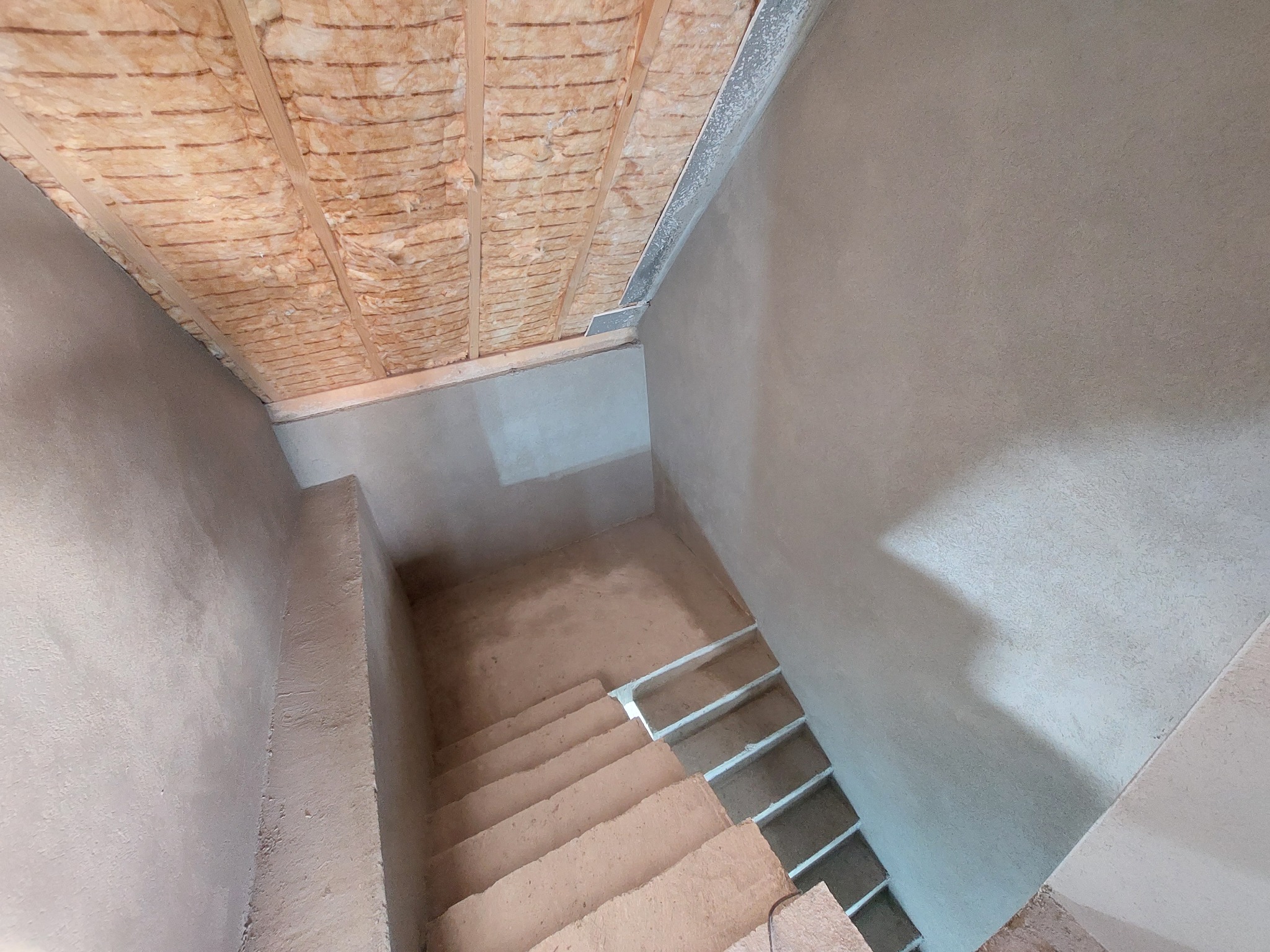 Wnętrze w trakcie tynkowania maszynowego, widoczne ocieplenie stropu wełną mineralną, surowe schody i świeżo otynkowane ściany w szarym odcieniu.